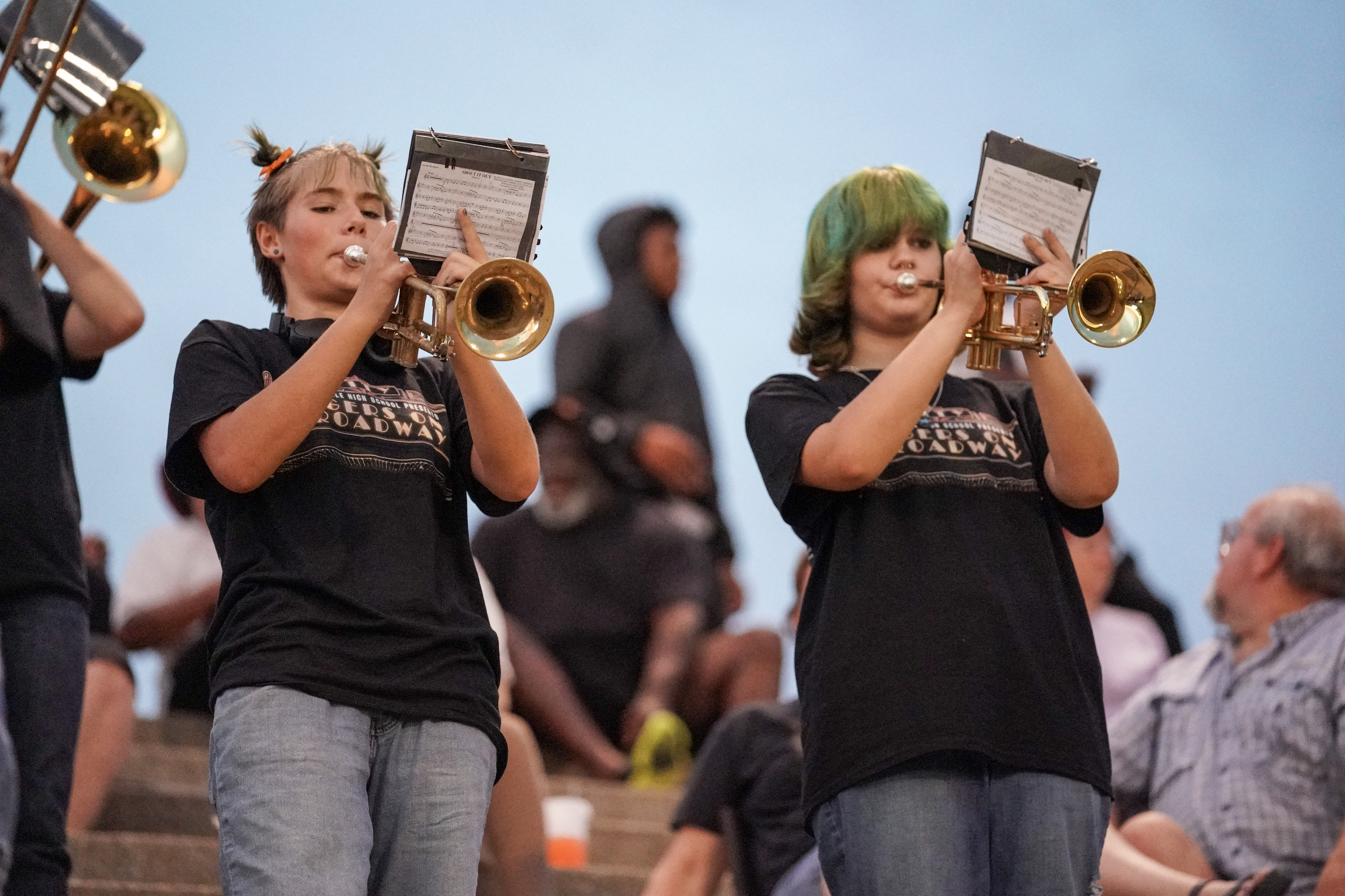 Two teenagers play trumpets on outdoor stadium steps, wearing matching black shirts and jeans. People sit in the background, some watching the performance, while the sky appears bright behind them.