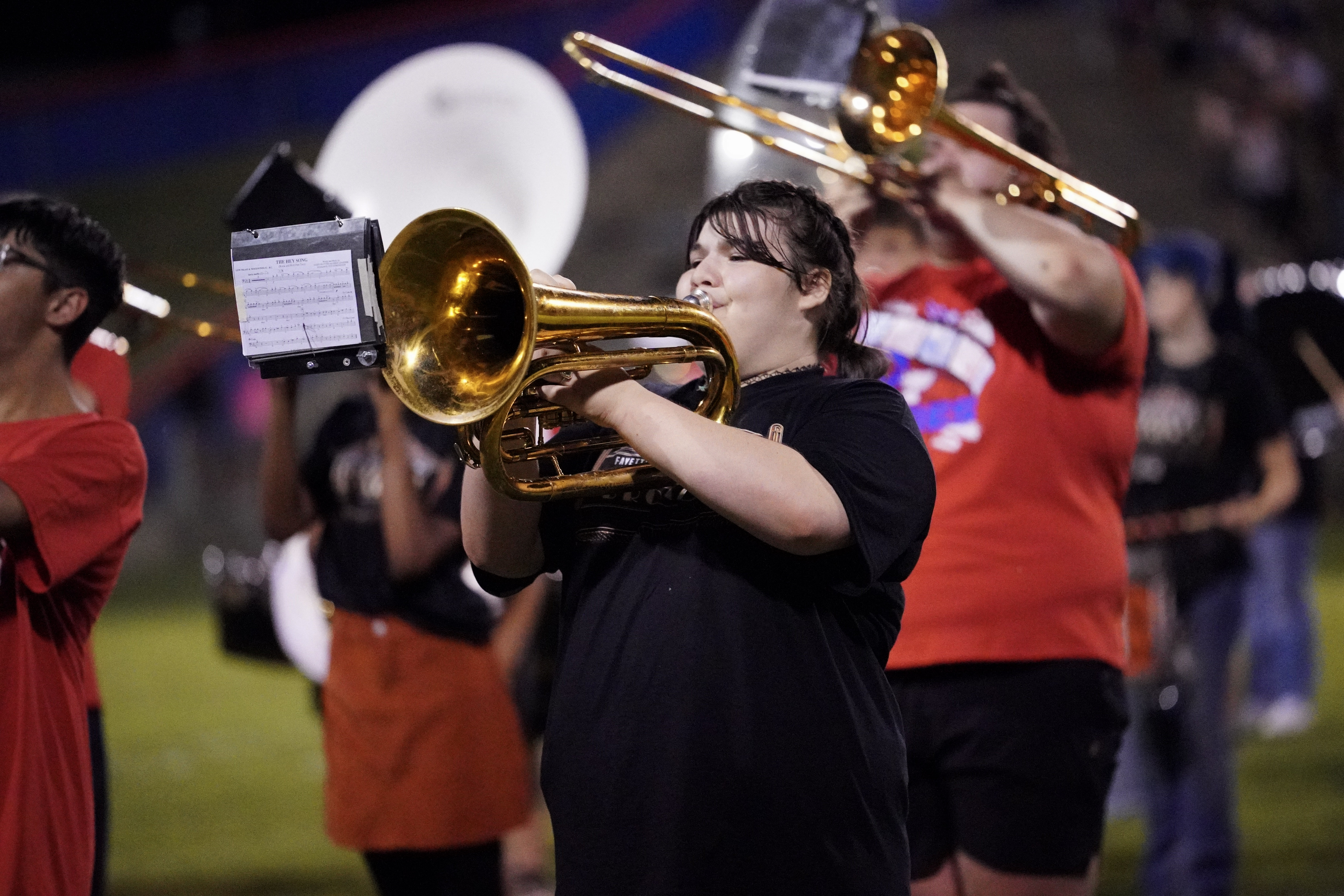 A marching band member plays a brass instrument on a field at night, surrounded by other musicians in casual clothing. The scene is energetic, with instruments gleaming under stadium lights.