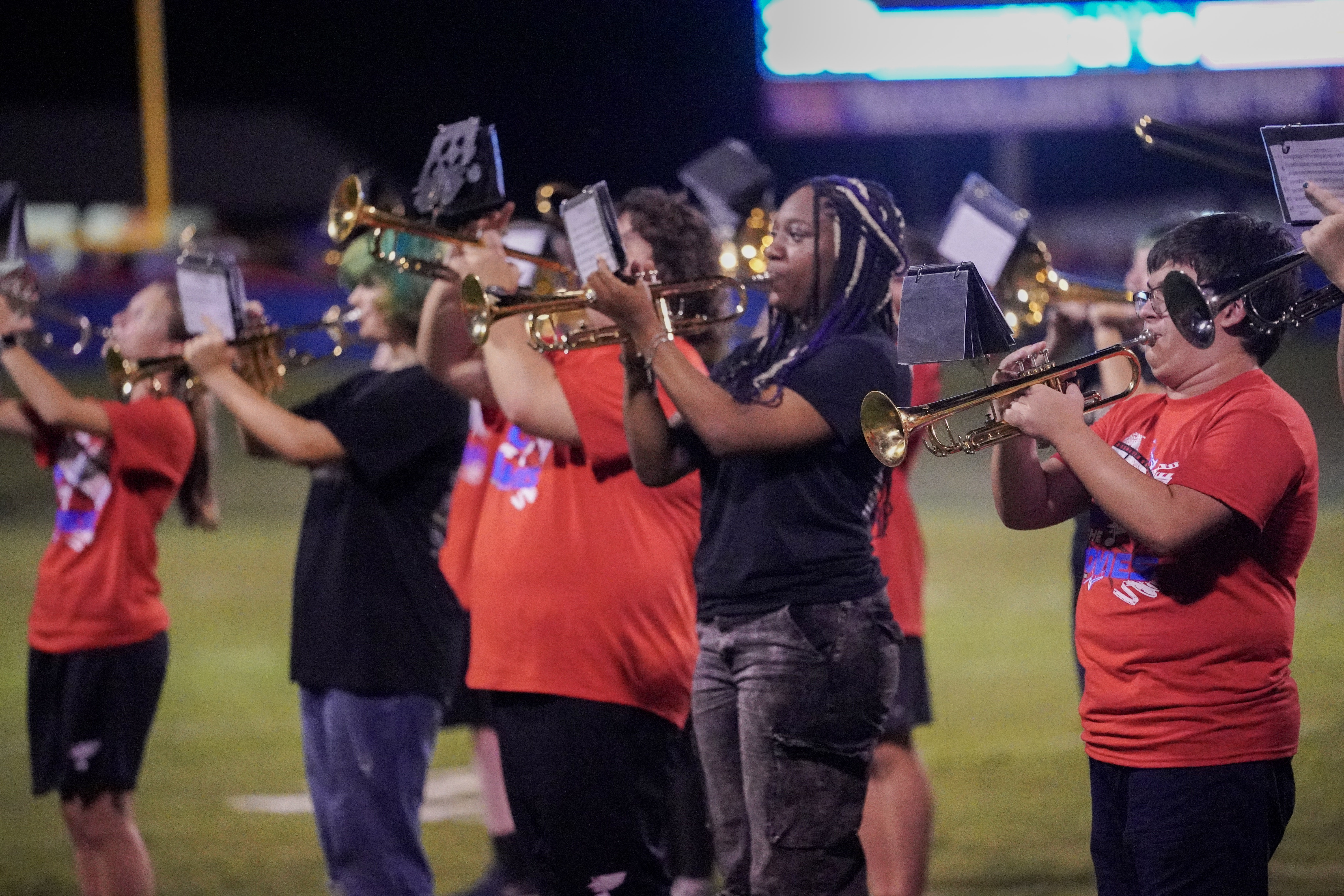 A group of young musicians play trumpets in formation on a field at night, some wearing red shirts and others in black, with sheet music attached to their instruments.