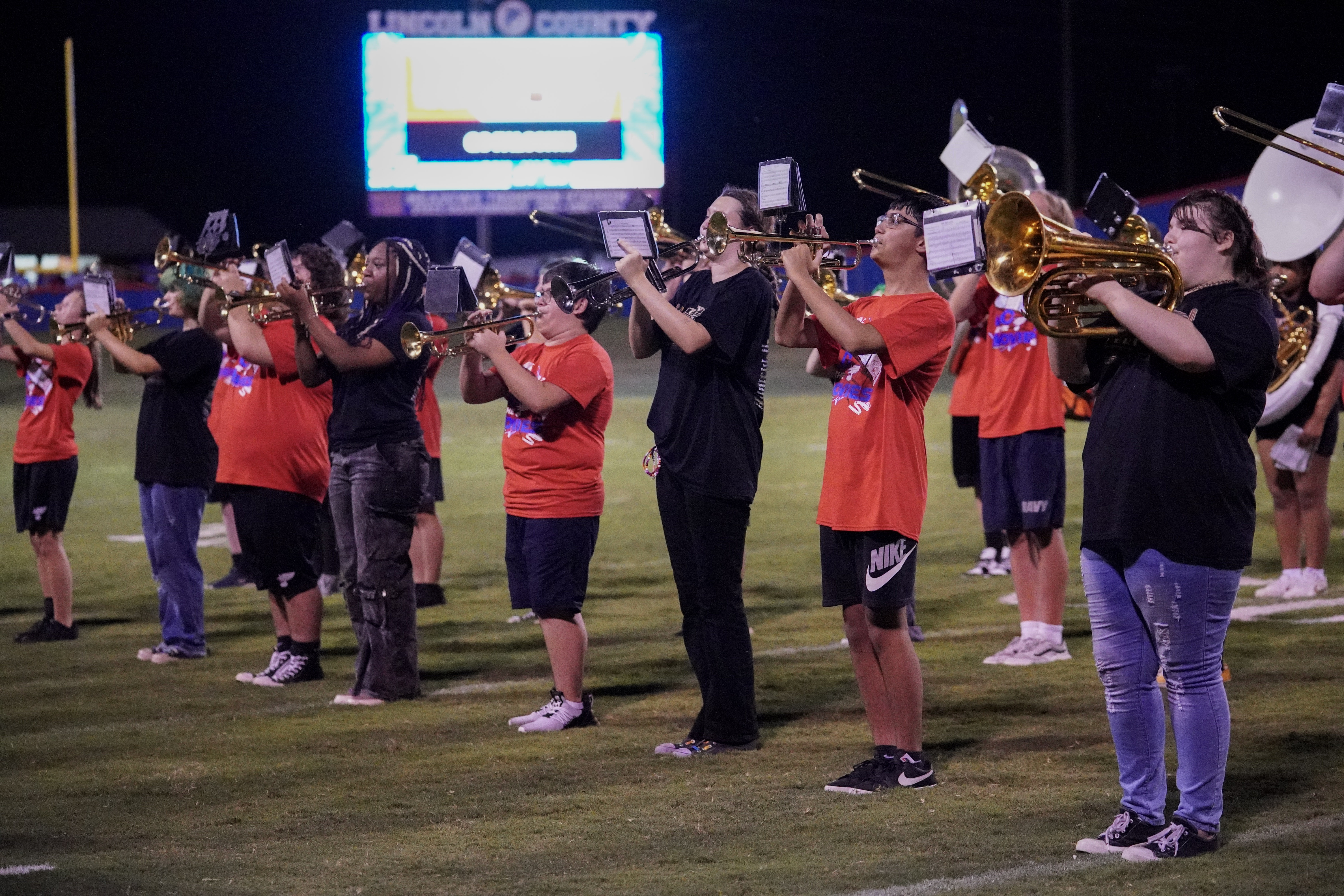 A marching band stands in formation on a football field at night, playing brass instruments. Some members wear red shirts, while others wear black, with a large scoreboard lit up in the background.