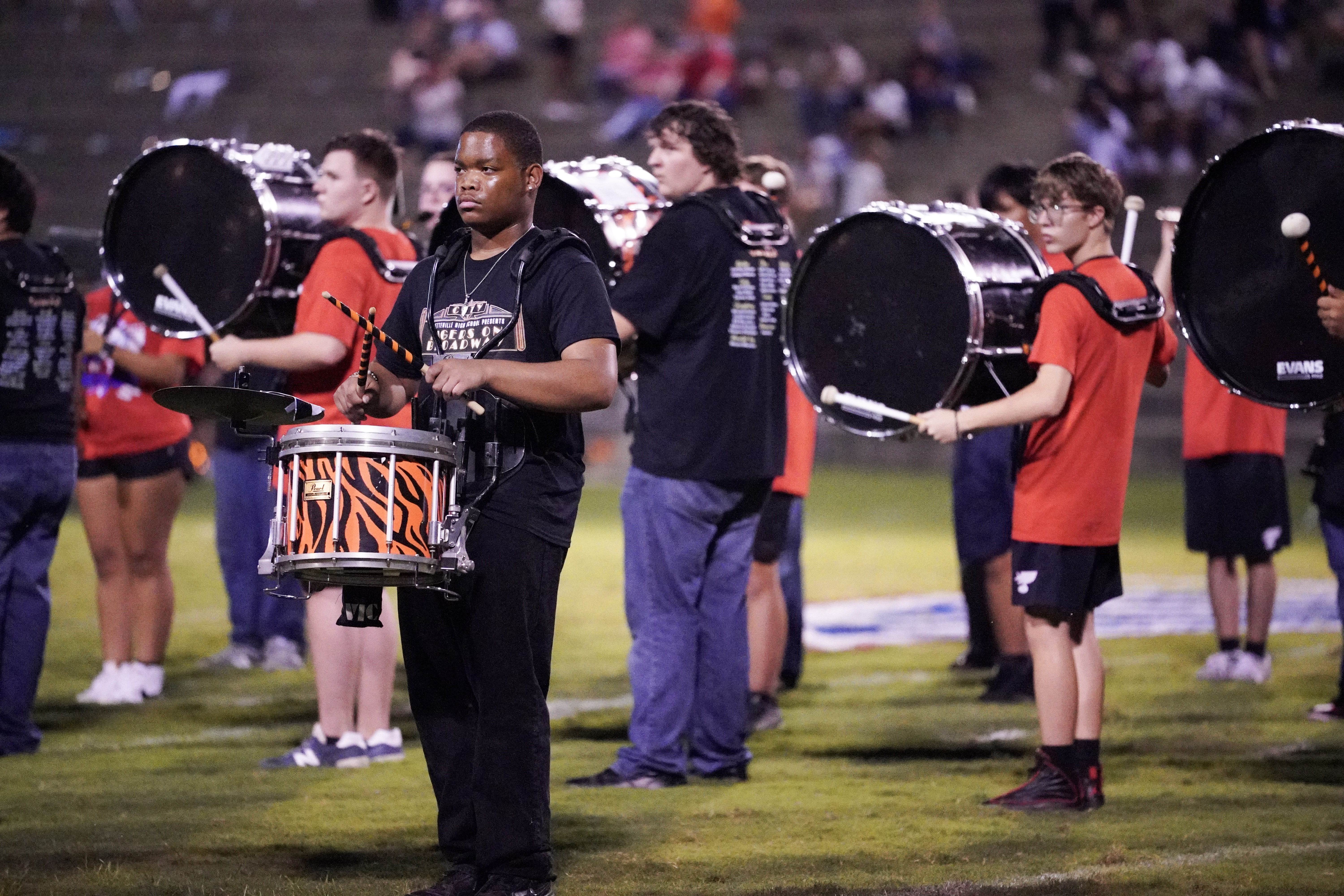 A marching band drumline performs on a football field at night, with several members playing snare and bass drums. The musicians wear black and red shirts, and the stands are filled with blurred spectators in the background.