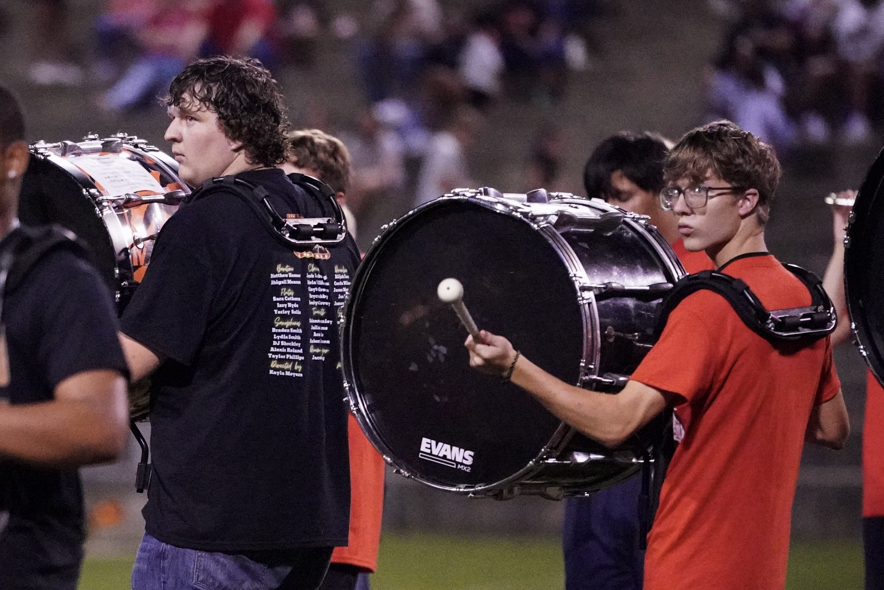 Two teenage boys stand side by side playing large bass drums at an outdoor event, possibly a marching band performance, with blurred spectators in the background. One boy looks ahead while the other glances toward the camera.
