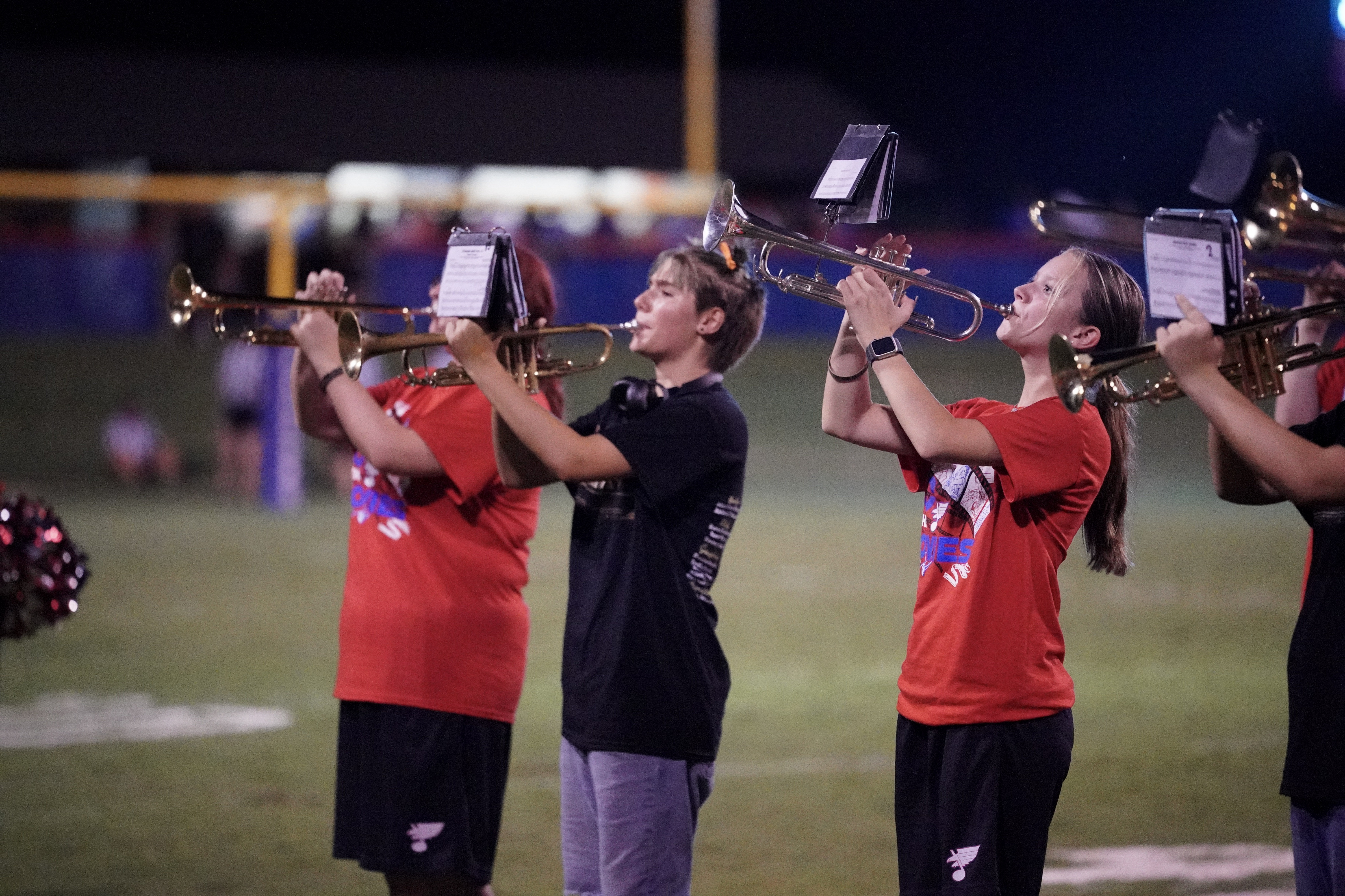 Three young people stand in a row on a football field at night, two wearing red shirts and playing trumpets, while the person in the middle, in black, stands with hands at their sides, looking forward.