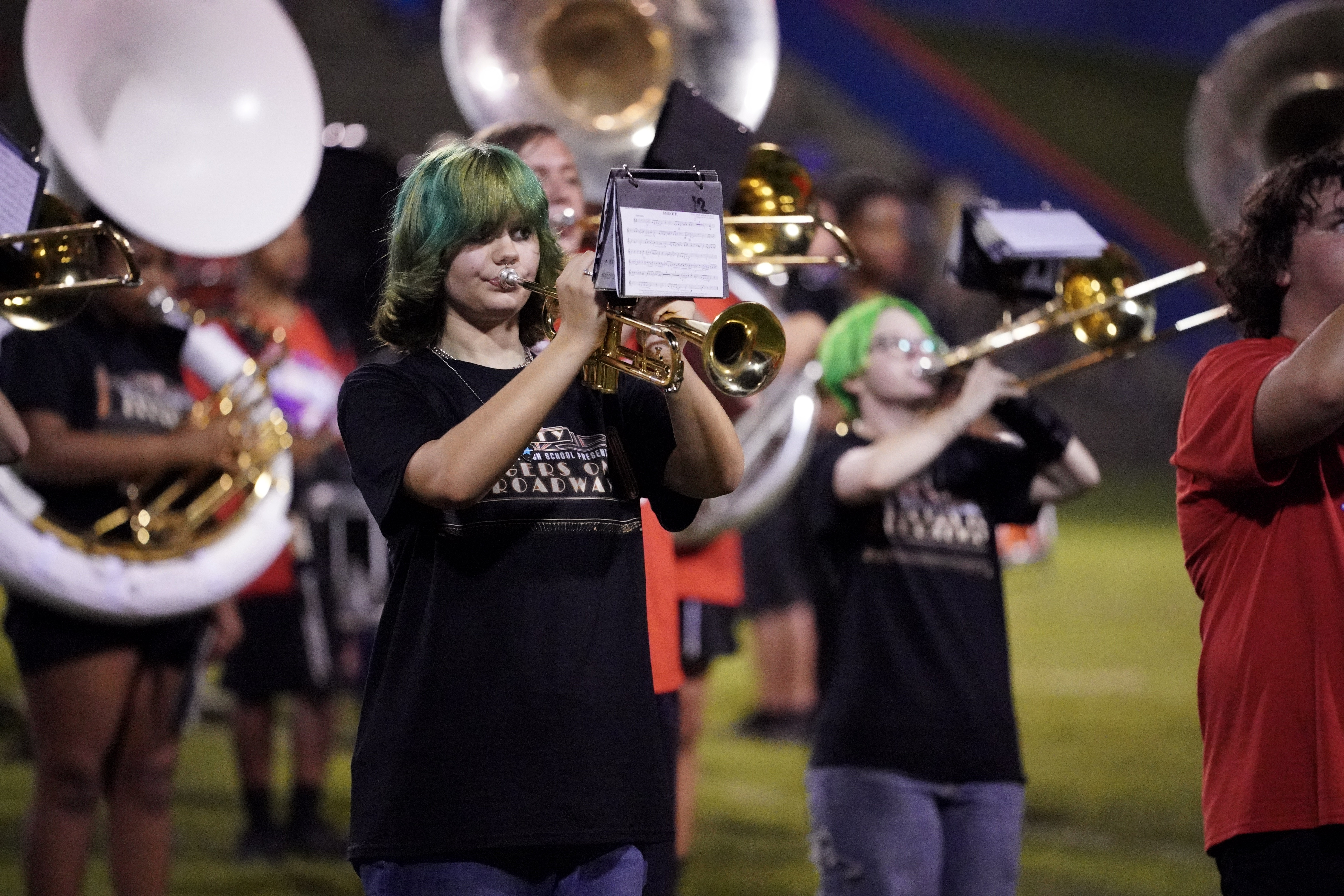 A marching band practices on a field at night. The focus is on a person with green hair playing a trumpet, wearing a black T-shirt. Other band members and brass instruments are visible in the background.