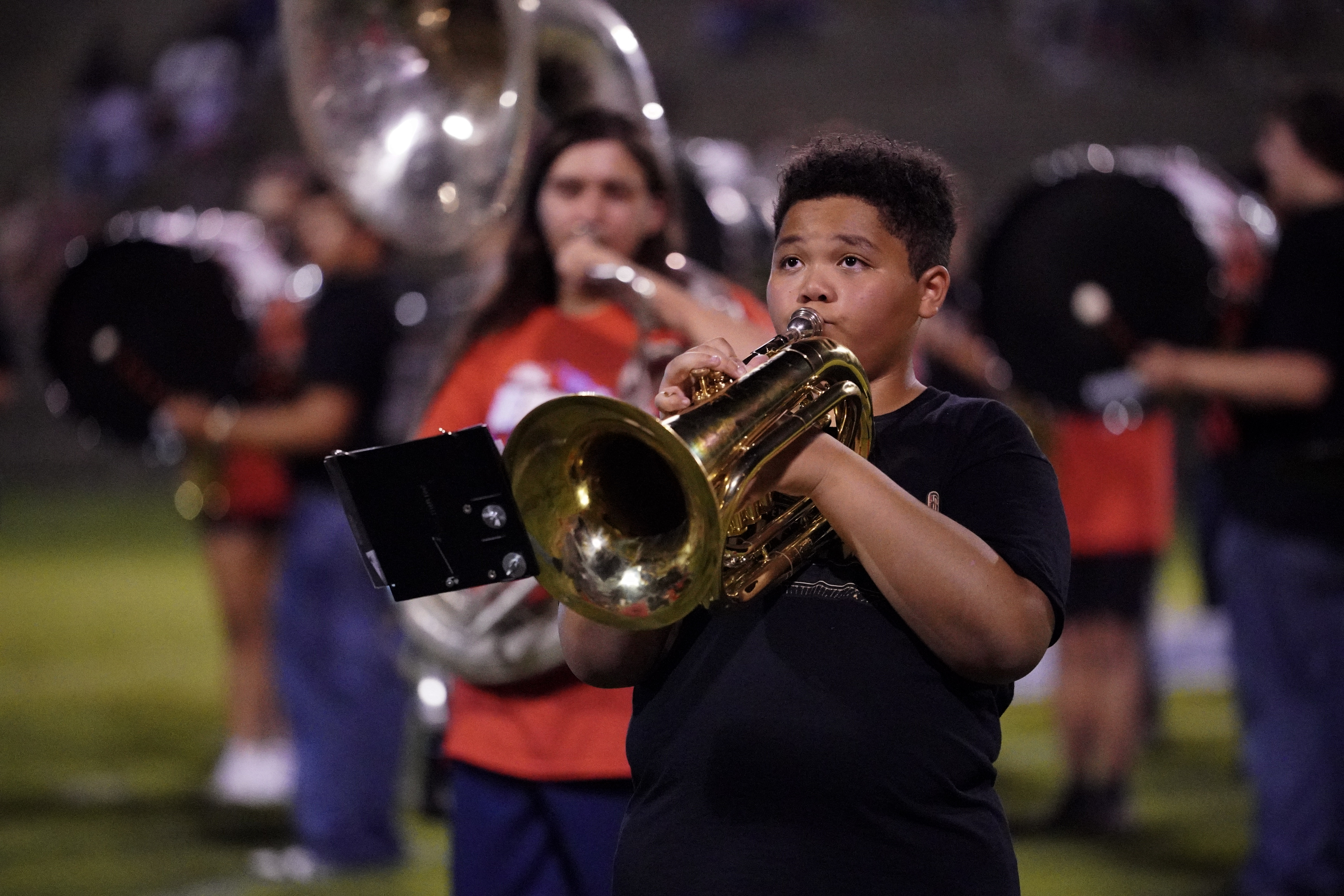A young boy plays a brass instrument in a marching band on a field, with other band members blurred in the background.