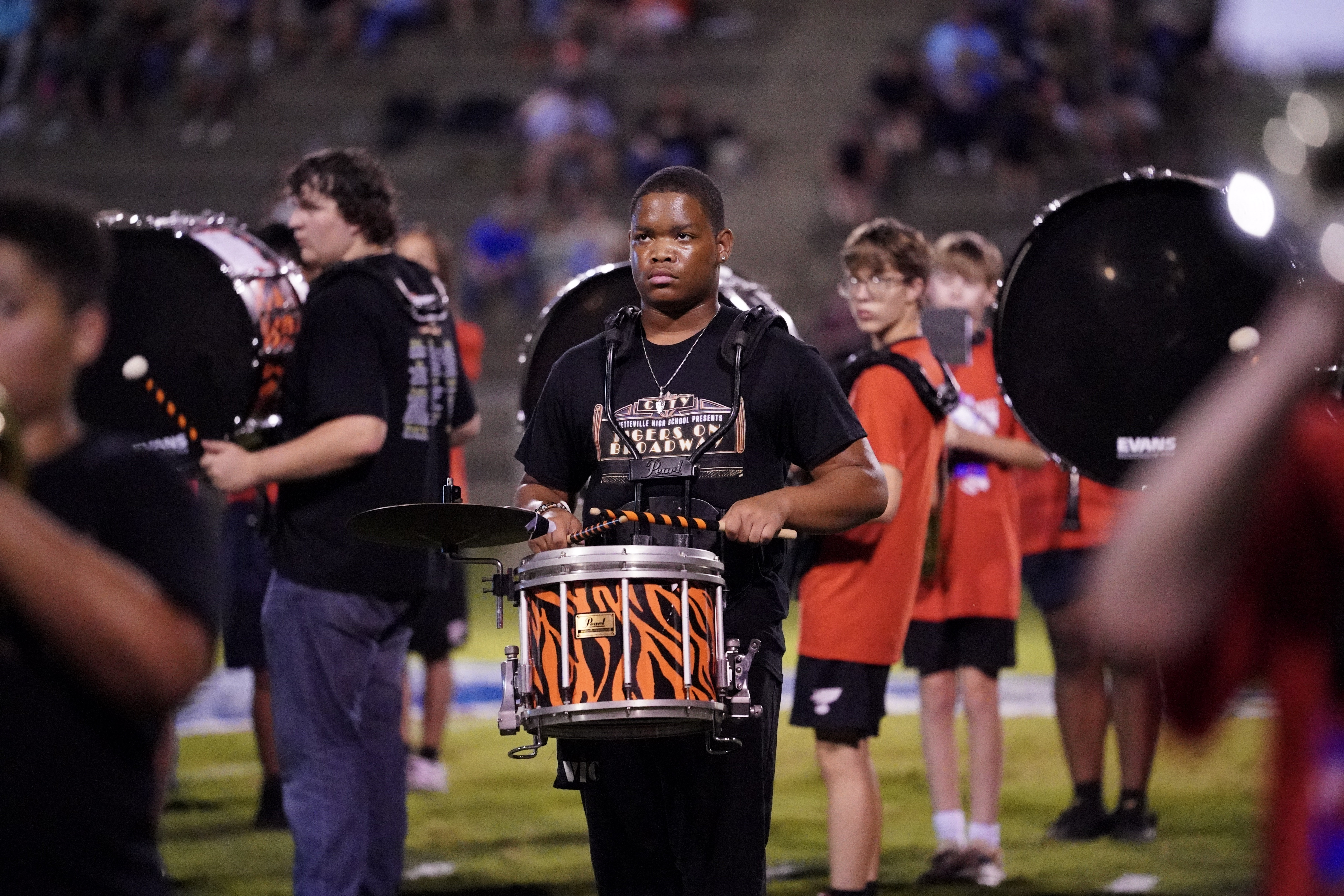 A focused young man in a black t-shirt plays a snare drum with an orange tiger-stripe design during a marching band performance on a field, surrounded by other drummers in black and red shirts.