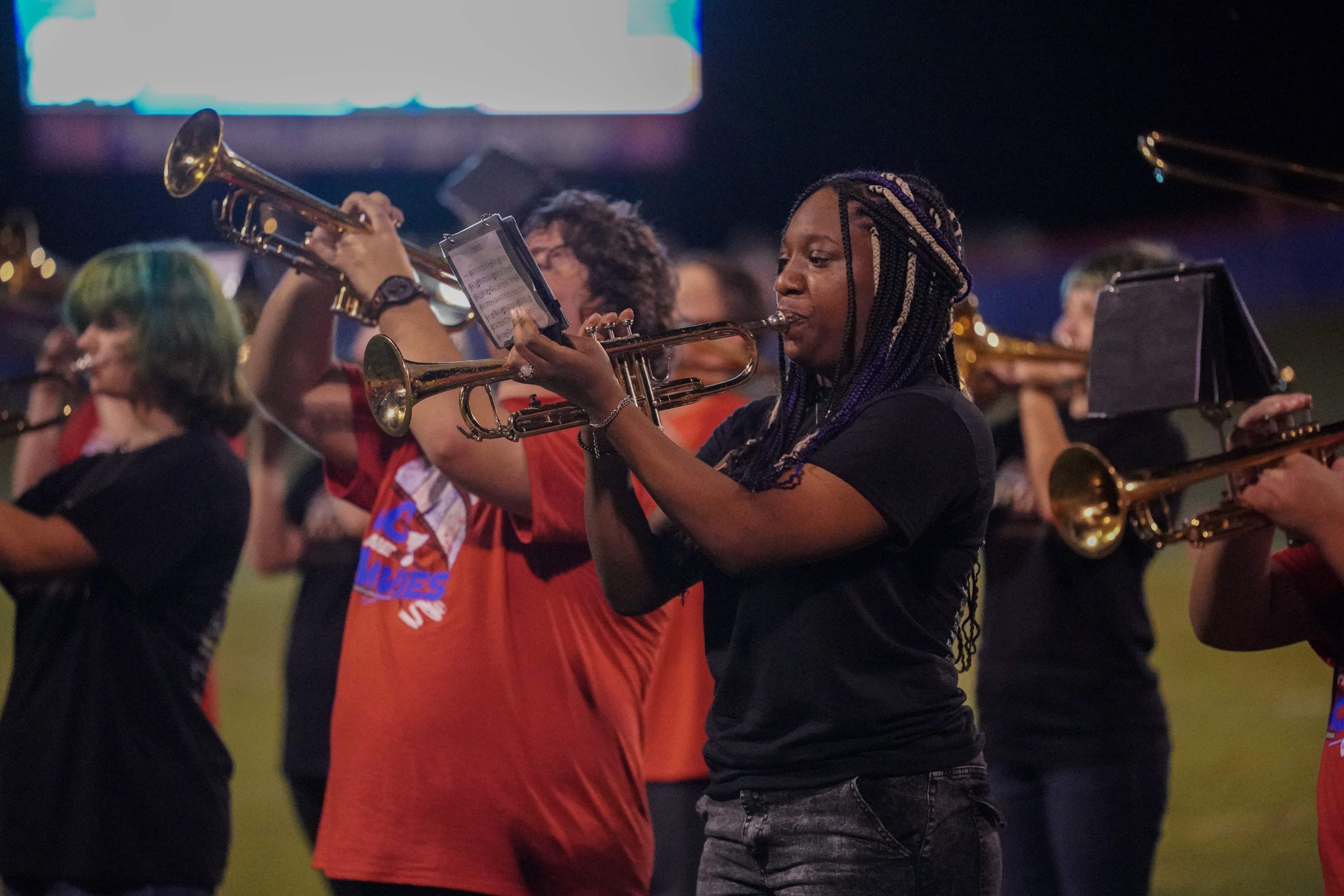 A group of marching band members play trumpets outdoors at night. The musicians wear casual clothes and focus on their instruments, with one woman in the foreground highlighted.
