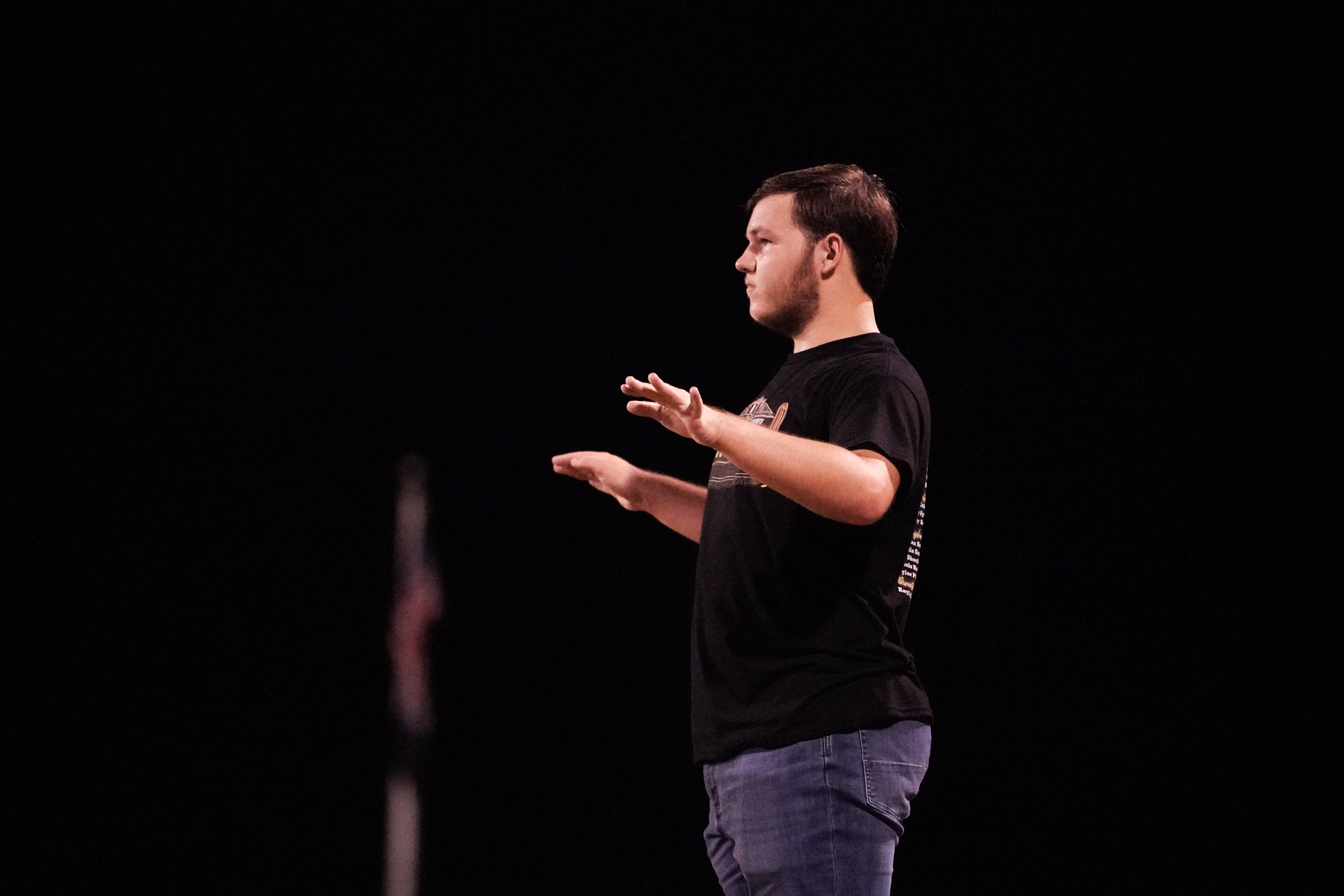 A person stands against a dark background with both arms raised and slightly bent, as if conducting or giving instructions. They wear a black T-shirt and blue jeans. A blurred flagpole is visible in the background.