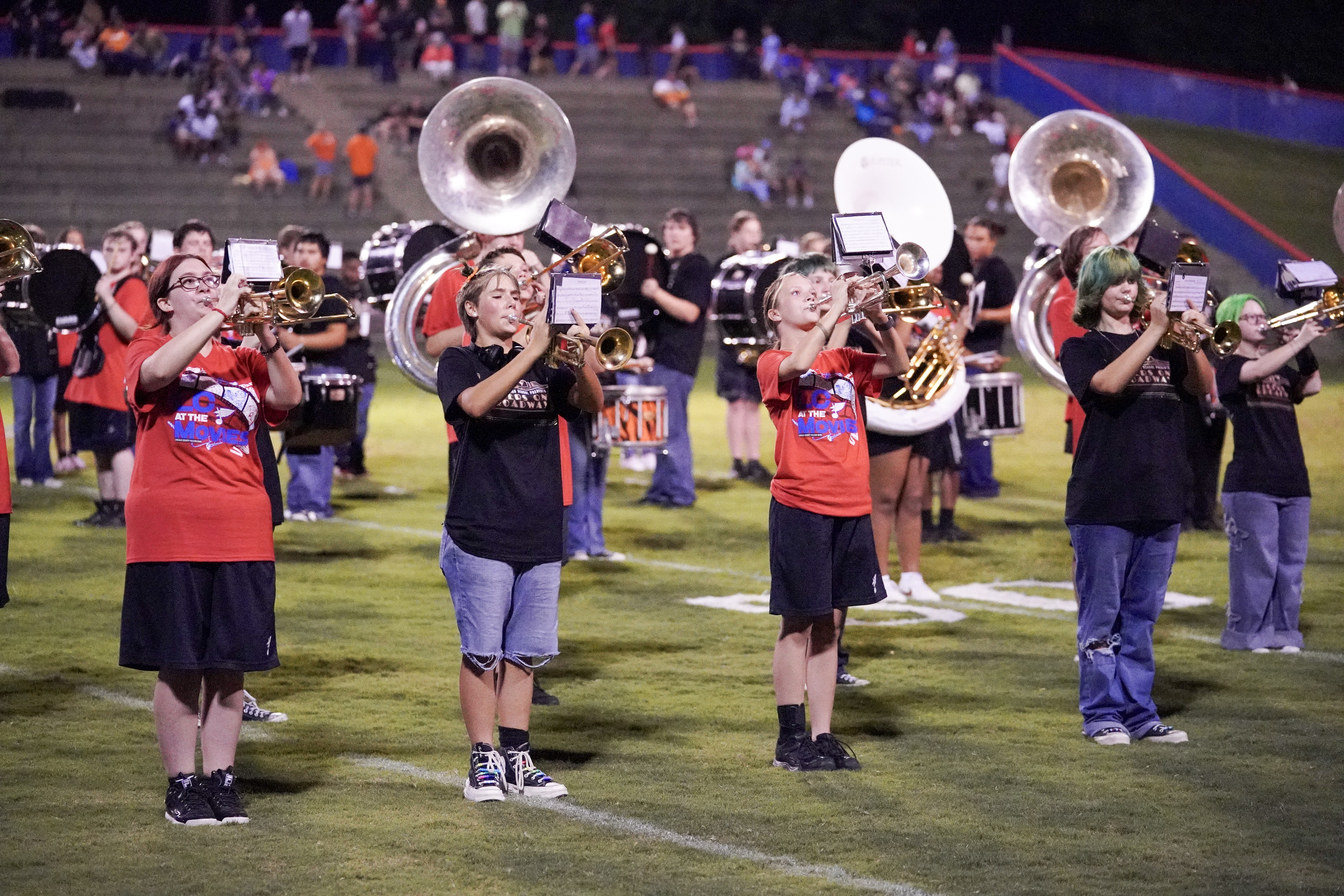 A group of marching band students play brass instruments on a football field at night, with people seated in bleachers in the background. The students wear red and black shirts.