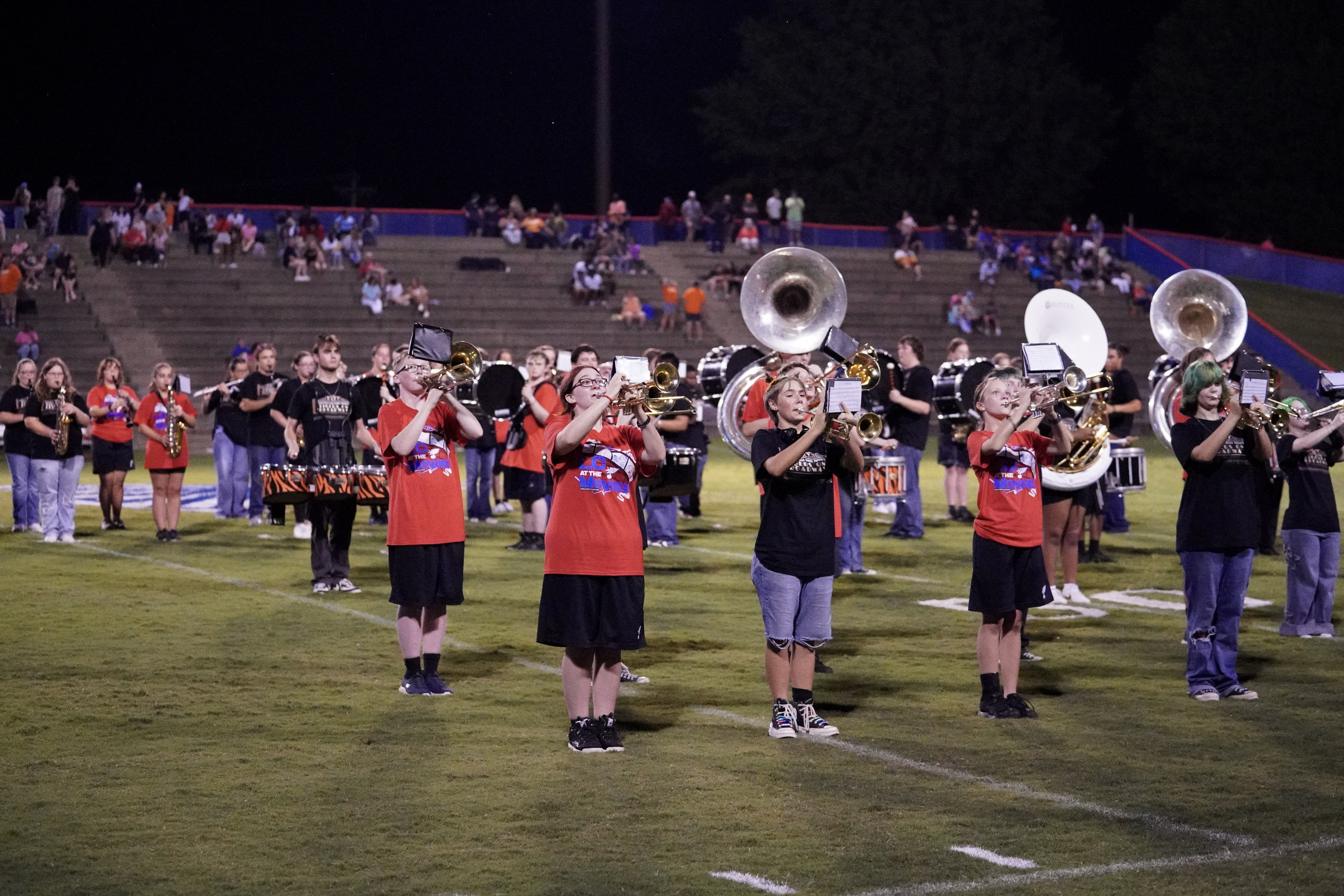 A marching band performs on a football field at night; band members play instruments while wearing uniforms, and an audience watches from the stadium bleachers in the background.