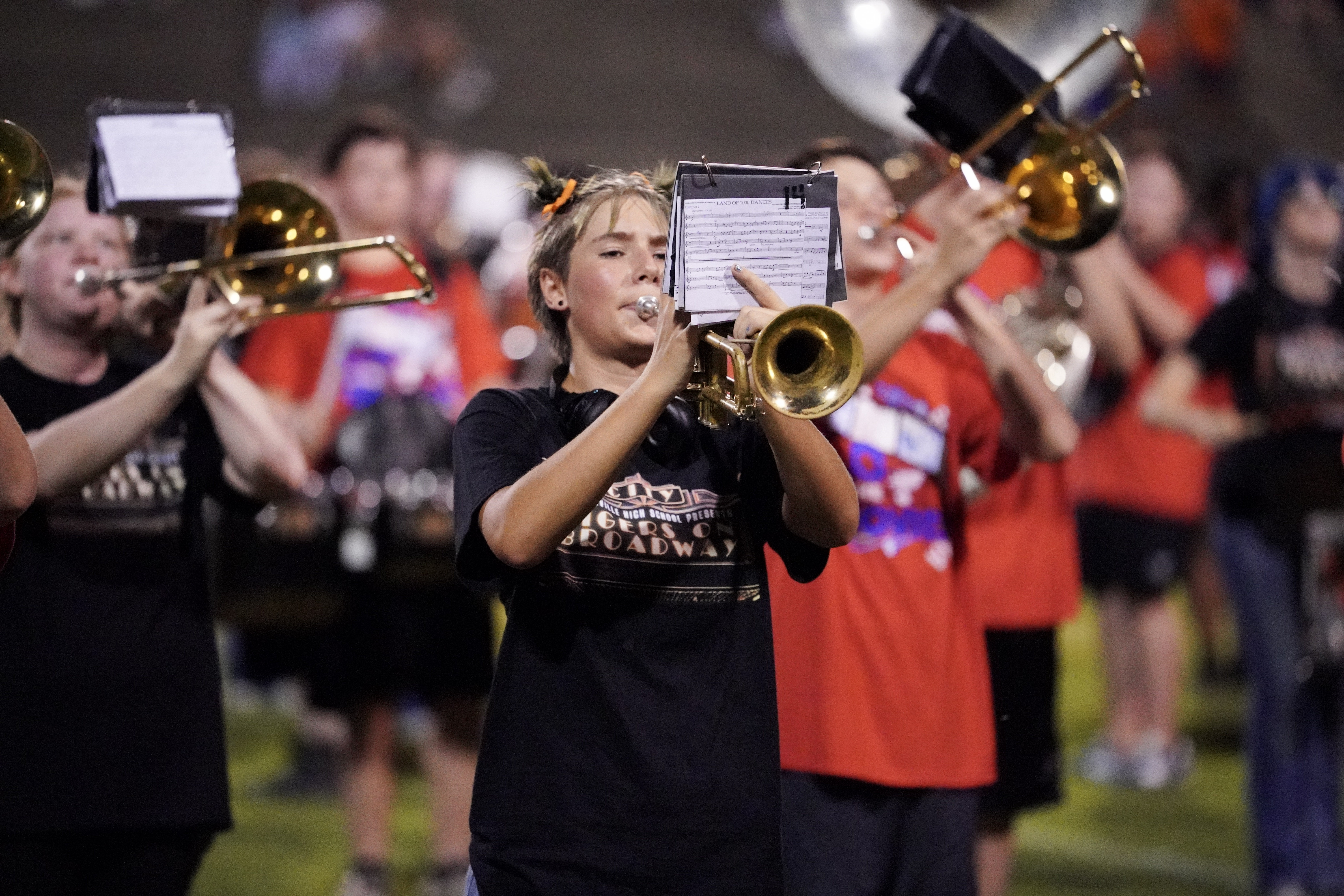 A marching band performs outdoors; a trumpet player in front plays while reading sheet music, with other band members in red and black shirts playing brass instruments behind them.