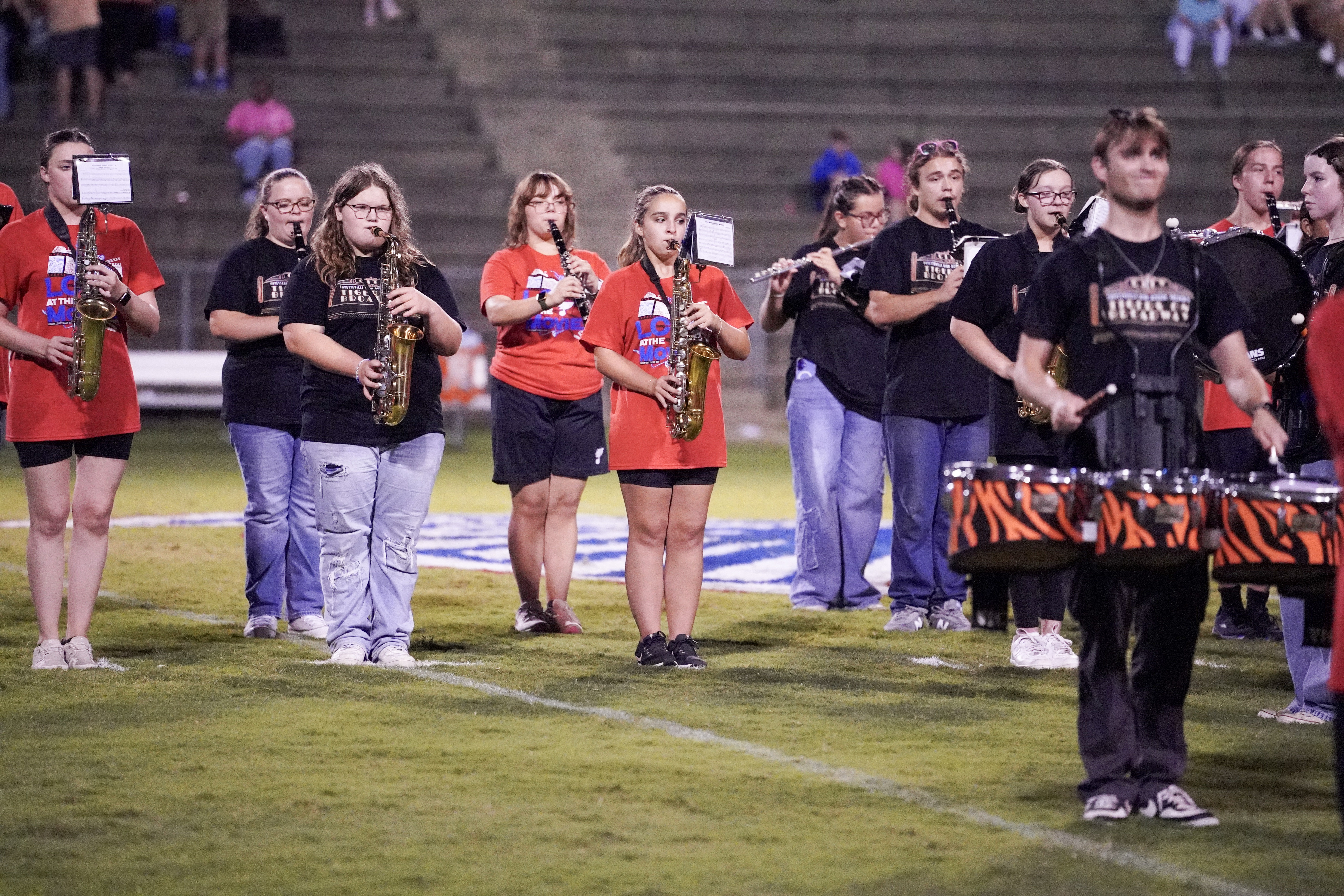 A group of marching band members, some in black shirts and some in red, play instruments on a field at night, with bleachers and a few spectators in the background.