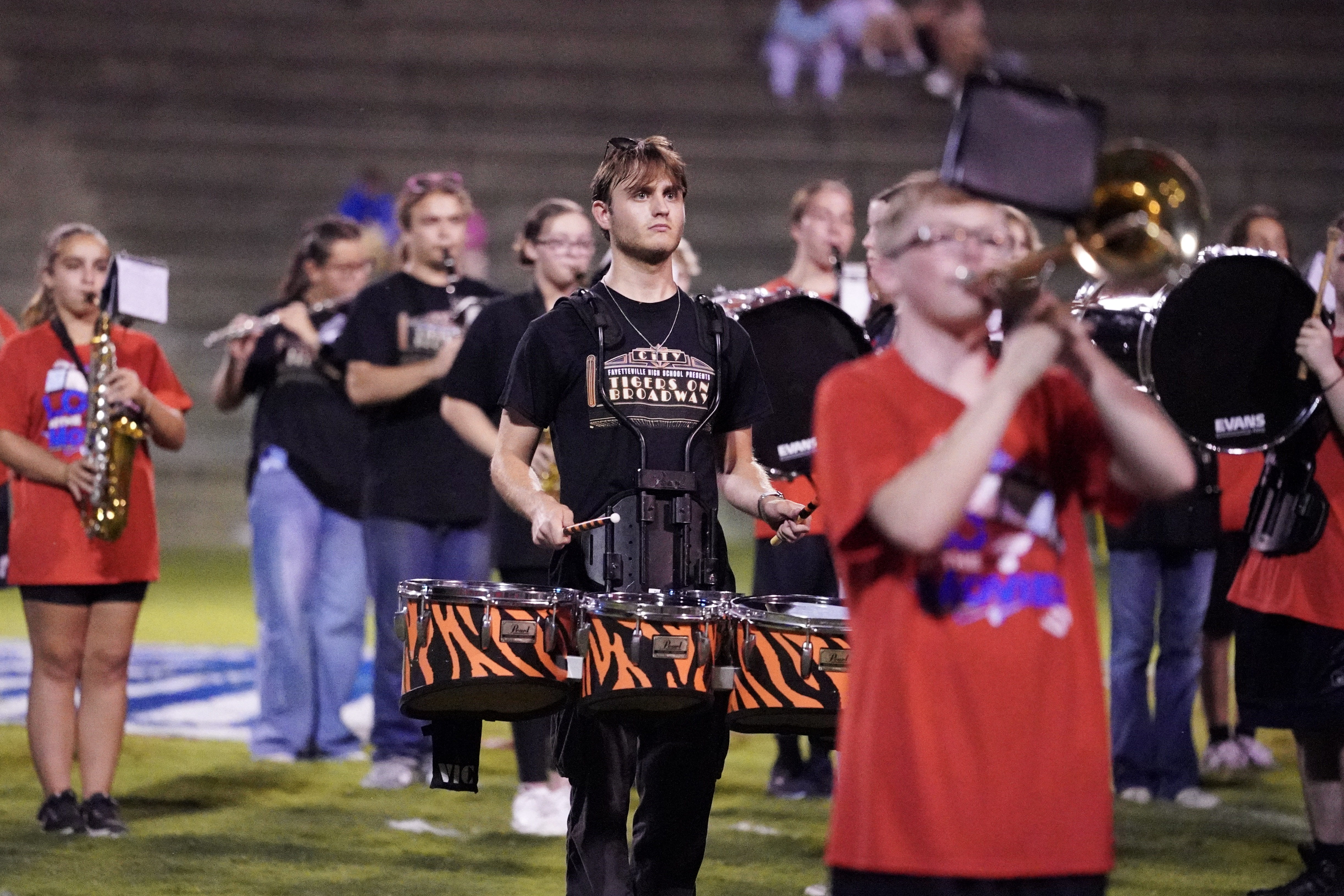 A high school marching band performs on a football field at night, with a drummer in the foreground wearing a black Tigers Go Seacats shirt and others playing various instruments in the background.
