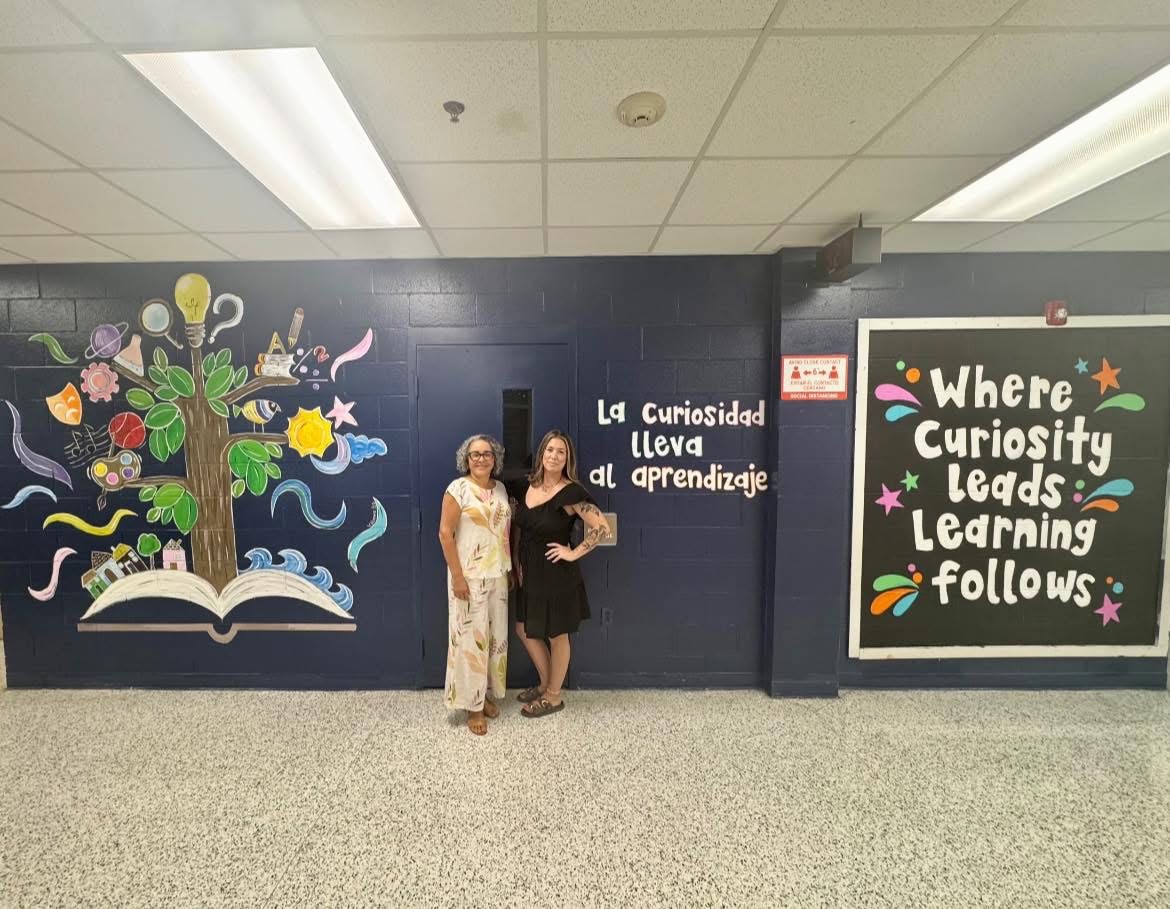 2 teachers standing in front of our school mural with theme of where curiosity leads learning follows