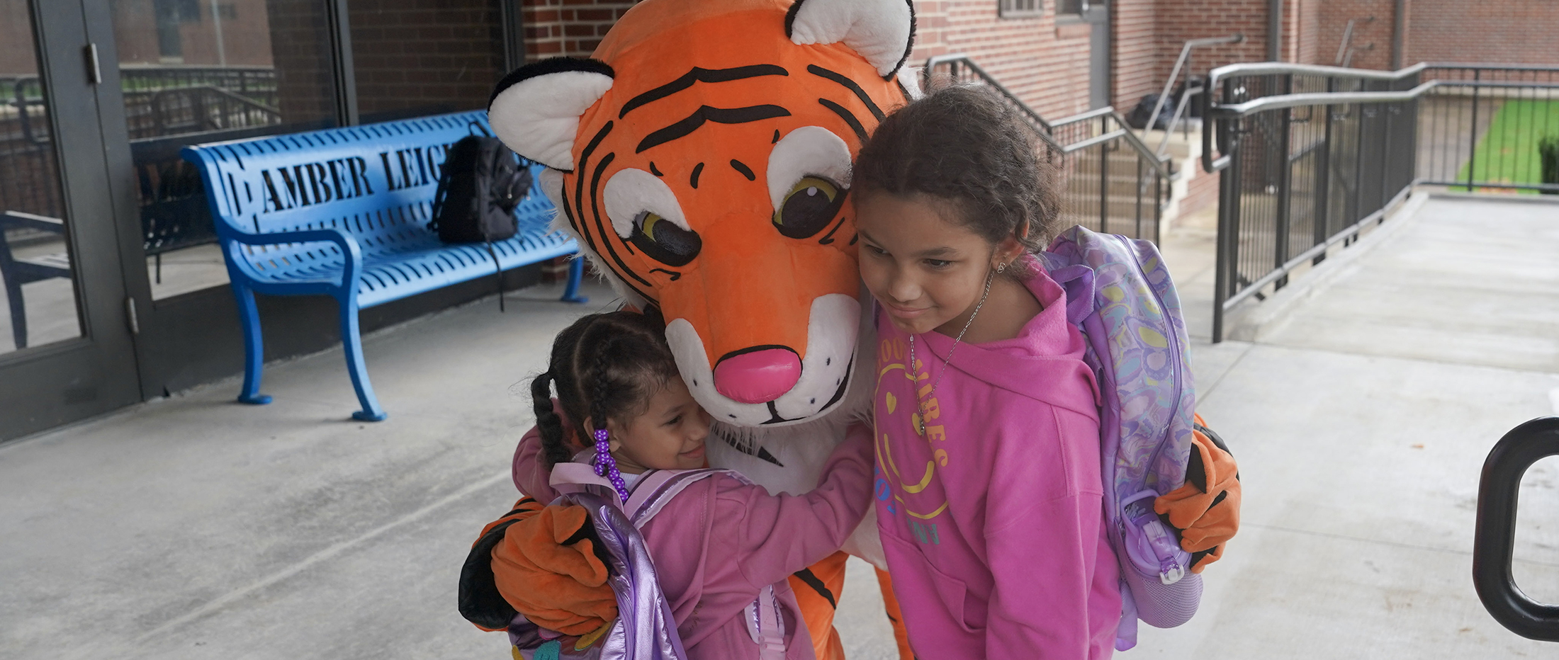 tiger mascot hugs two young students in front of a school