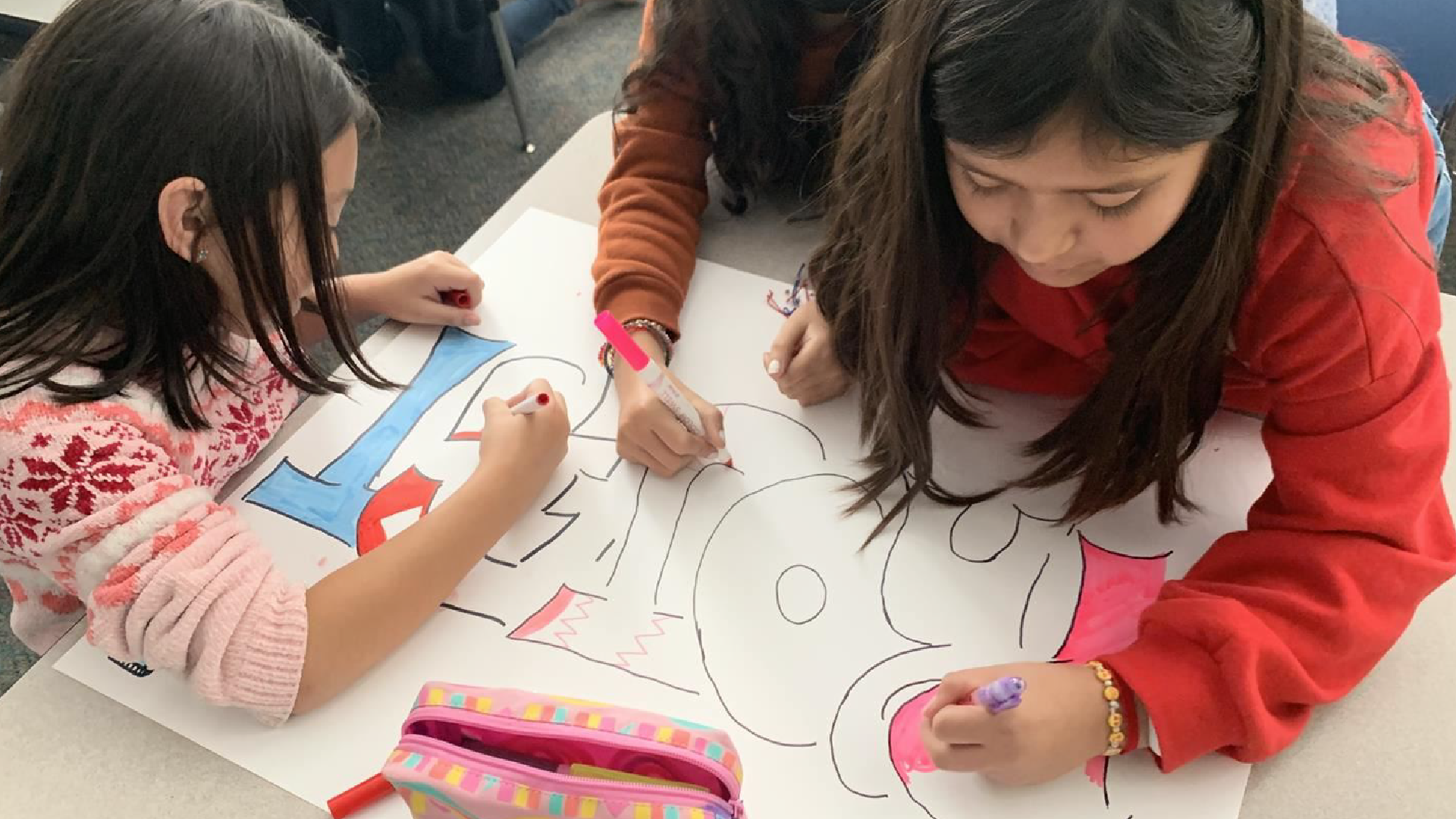 Two young girls in casual clothing are sitting at a table, intently coloring and drawing on a large sheet of paper in front of them. The background appears to be a classroom or educational setting, with other students and materials visible.