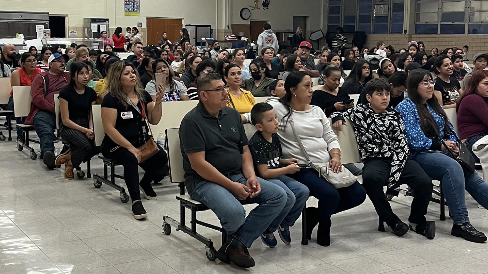 A large group of people, mostly young adults, are seated in a classroom or auditorium setting, with various individuals visible in the foreground and background.