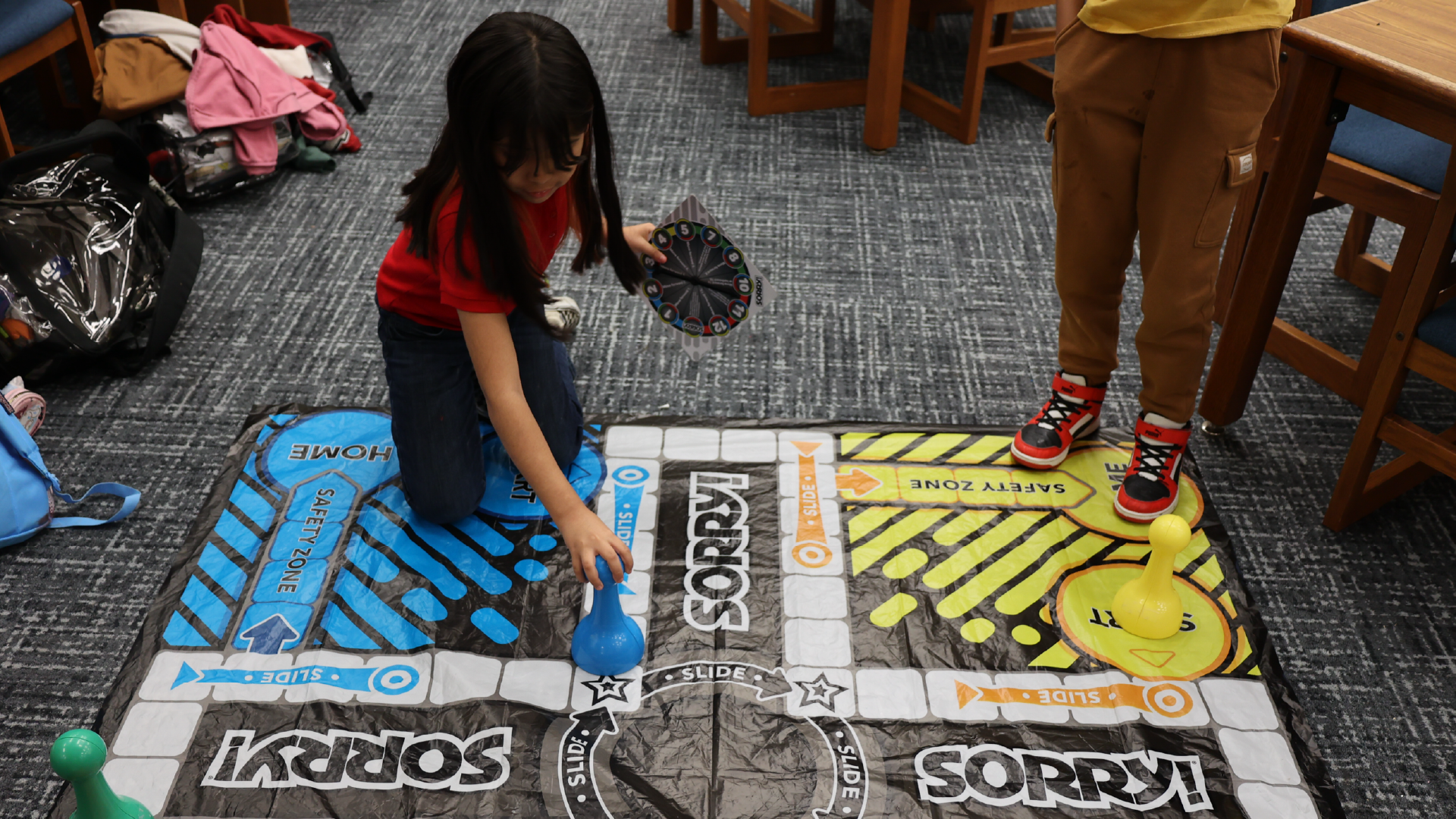 The image shows a cluttered floor with various items scattered around, including a backpack, clothing, and a colorful mat or rug with text and graphics.