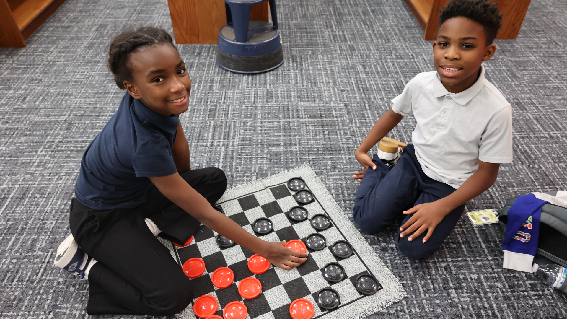 Two young boys are playing a game of checkers on the floor, surrounded by a tiled floor and what appears to be a classroom or educational setting.