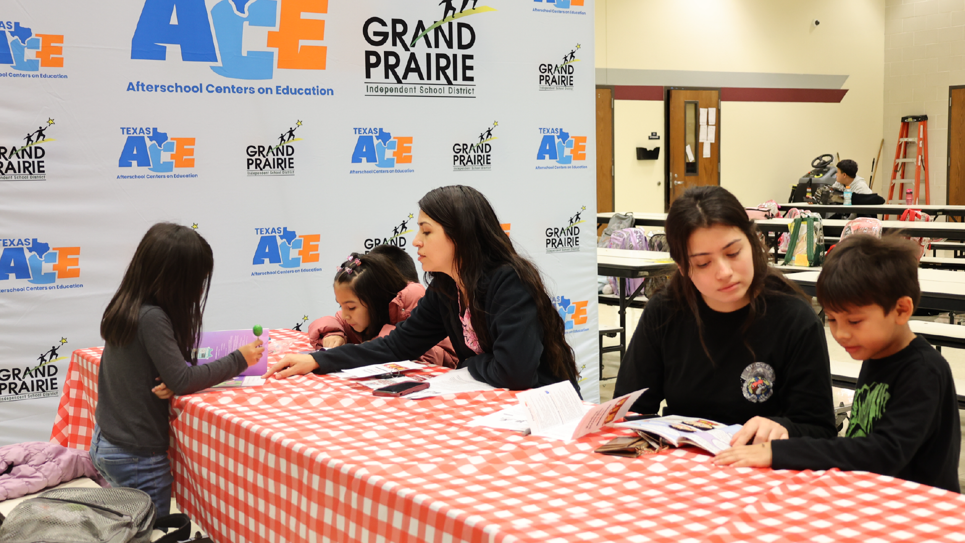 The image shows a group of people sitting at a table in what appears to be an educational or community event, with a backdrop featuring the logo and branding of "Grand Prairie".