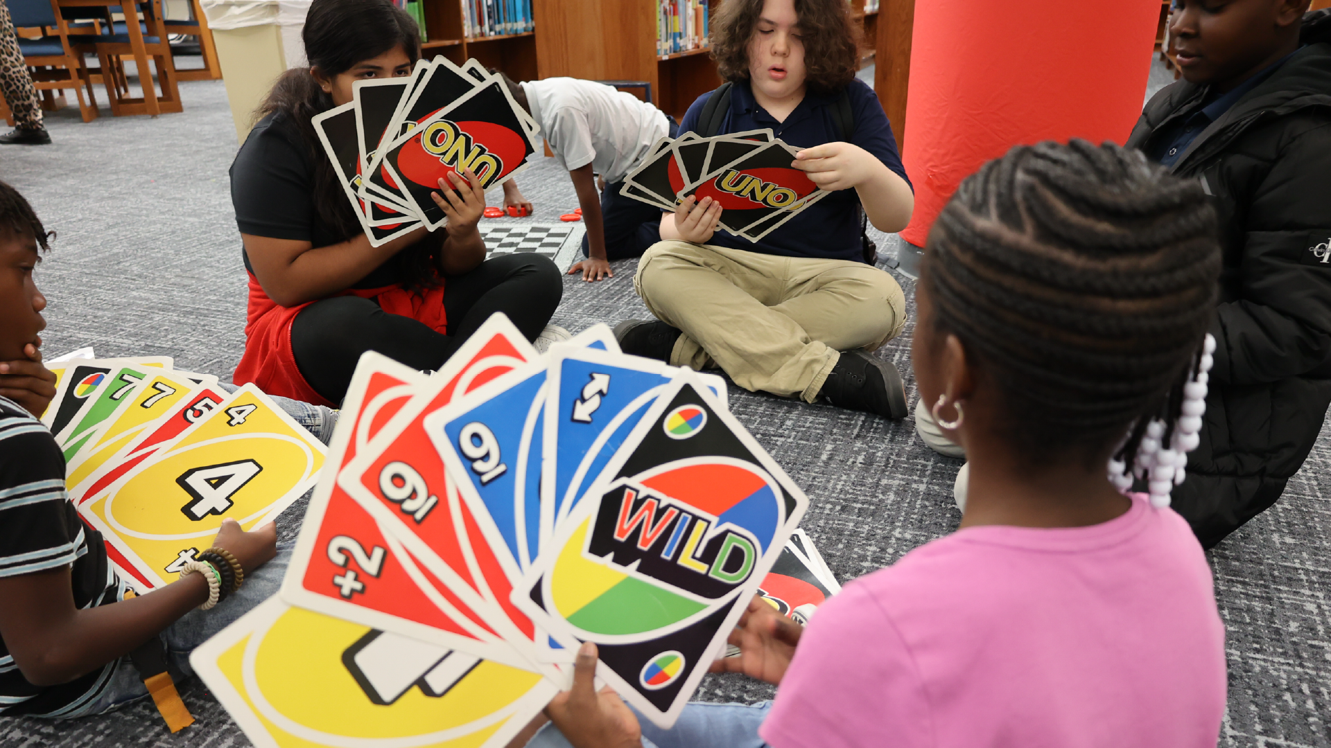 A group of people, including several young individuals, are gathered around a table playing a card game, with colorful cards and game pieces visible in the foreground.