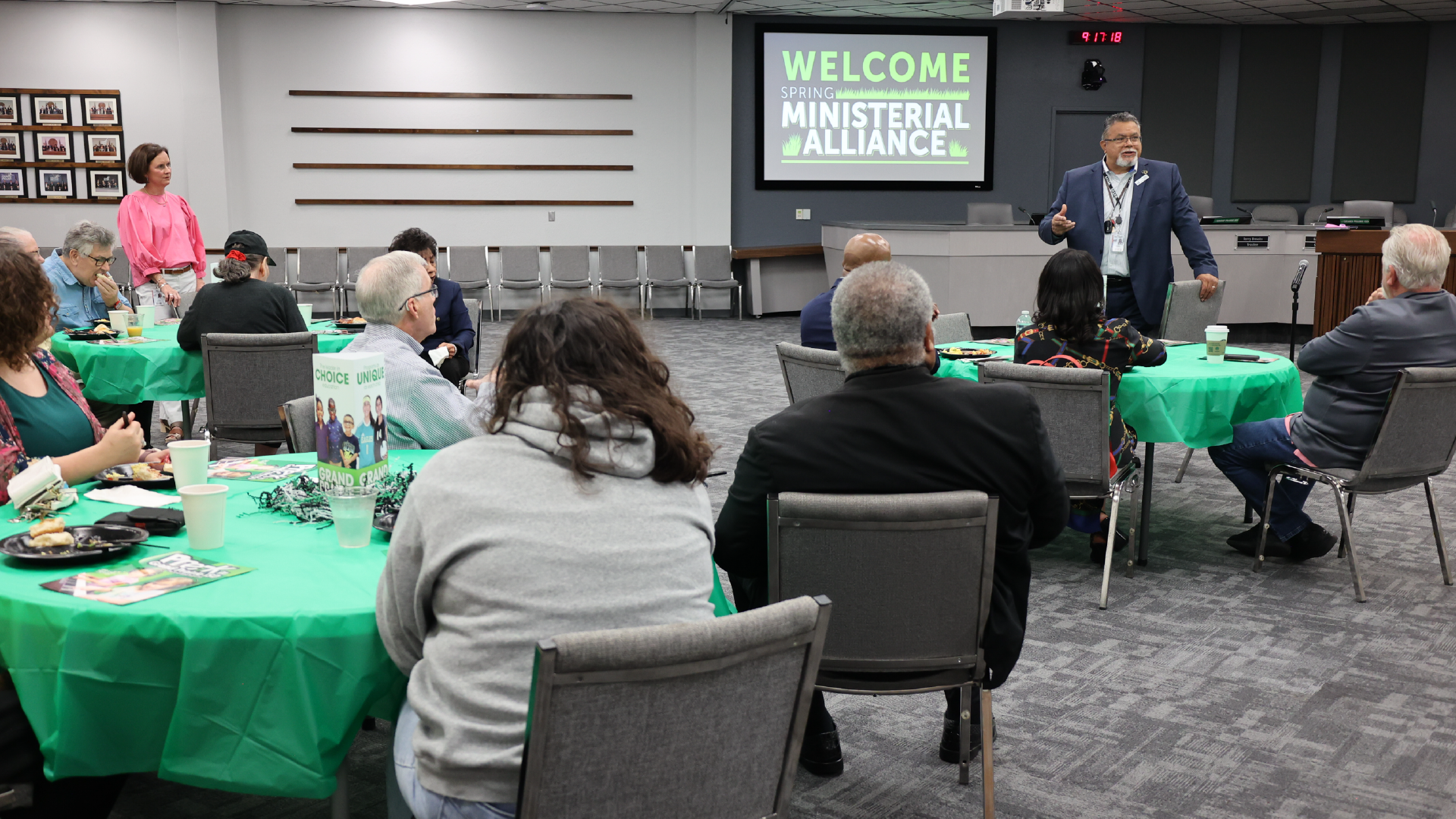 A group of people seated at tables in a conference room, with a large "Welcome" sign displayed on a screen in the background.