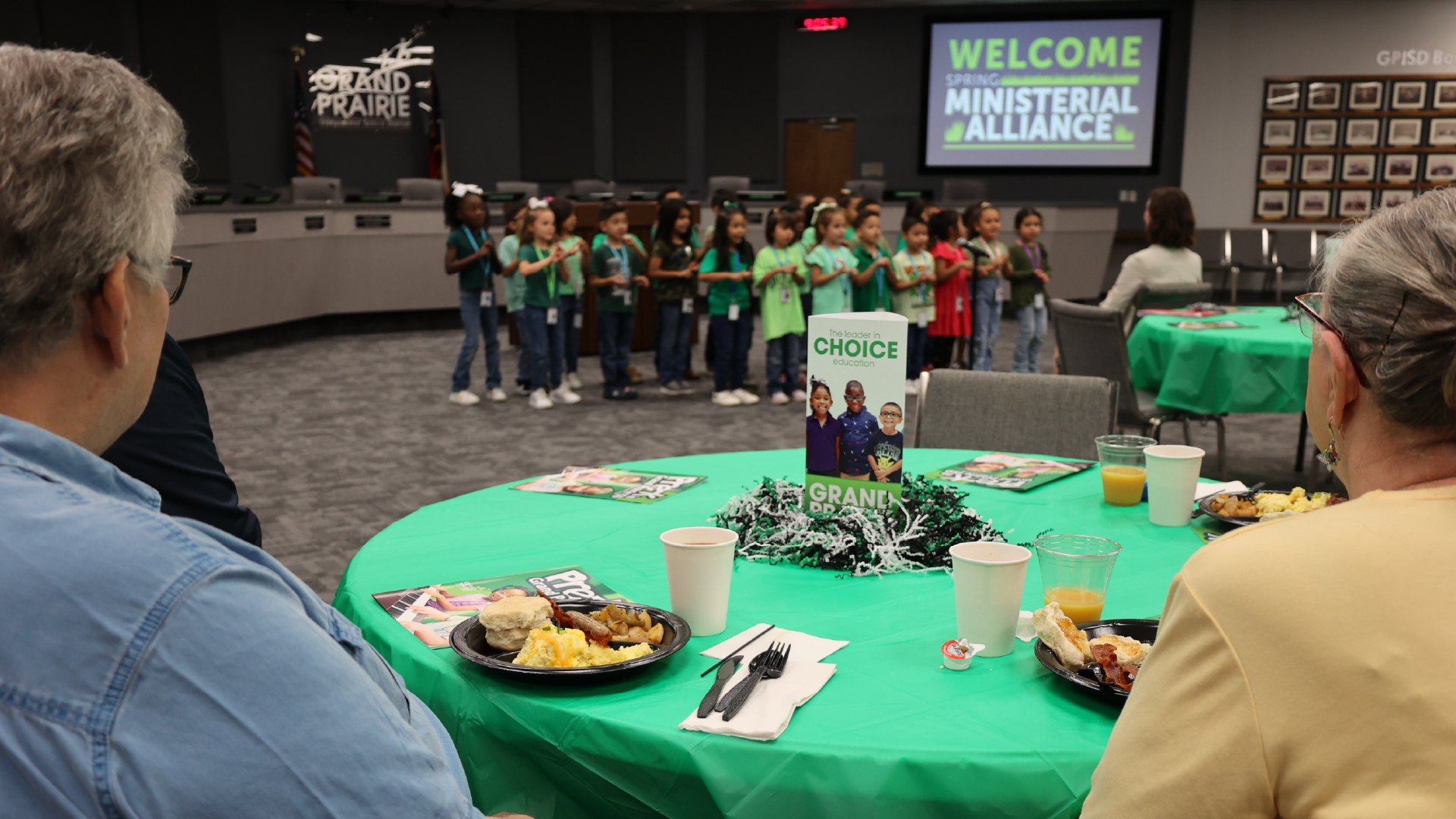The image shows a group of people gathered in a room, with a table in the foreground set up with various food items and a sign that says "Welcome Ministerial Alliance" displayed on a screen in the background.