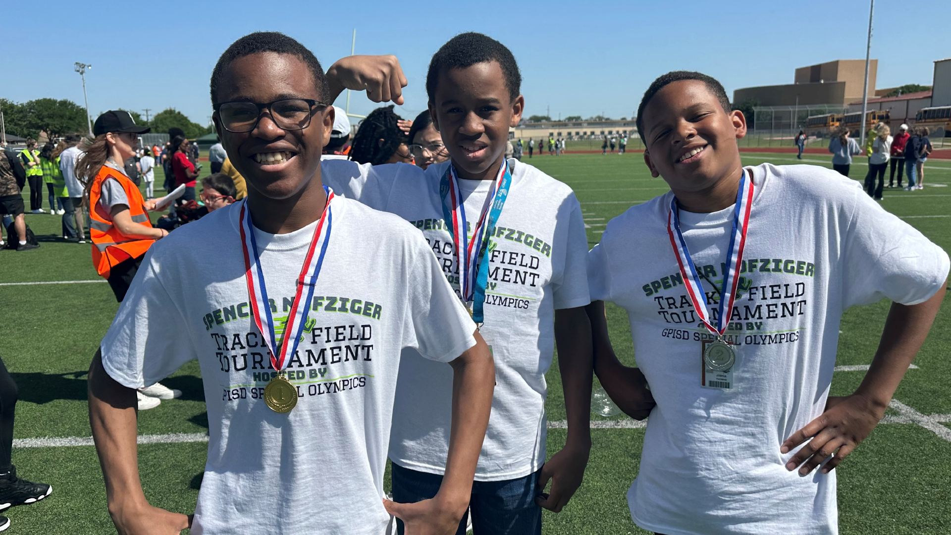 Three young men wearing white t-shirts with text on them are standing together on a sports field, with other people visible in the background.