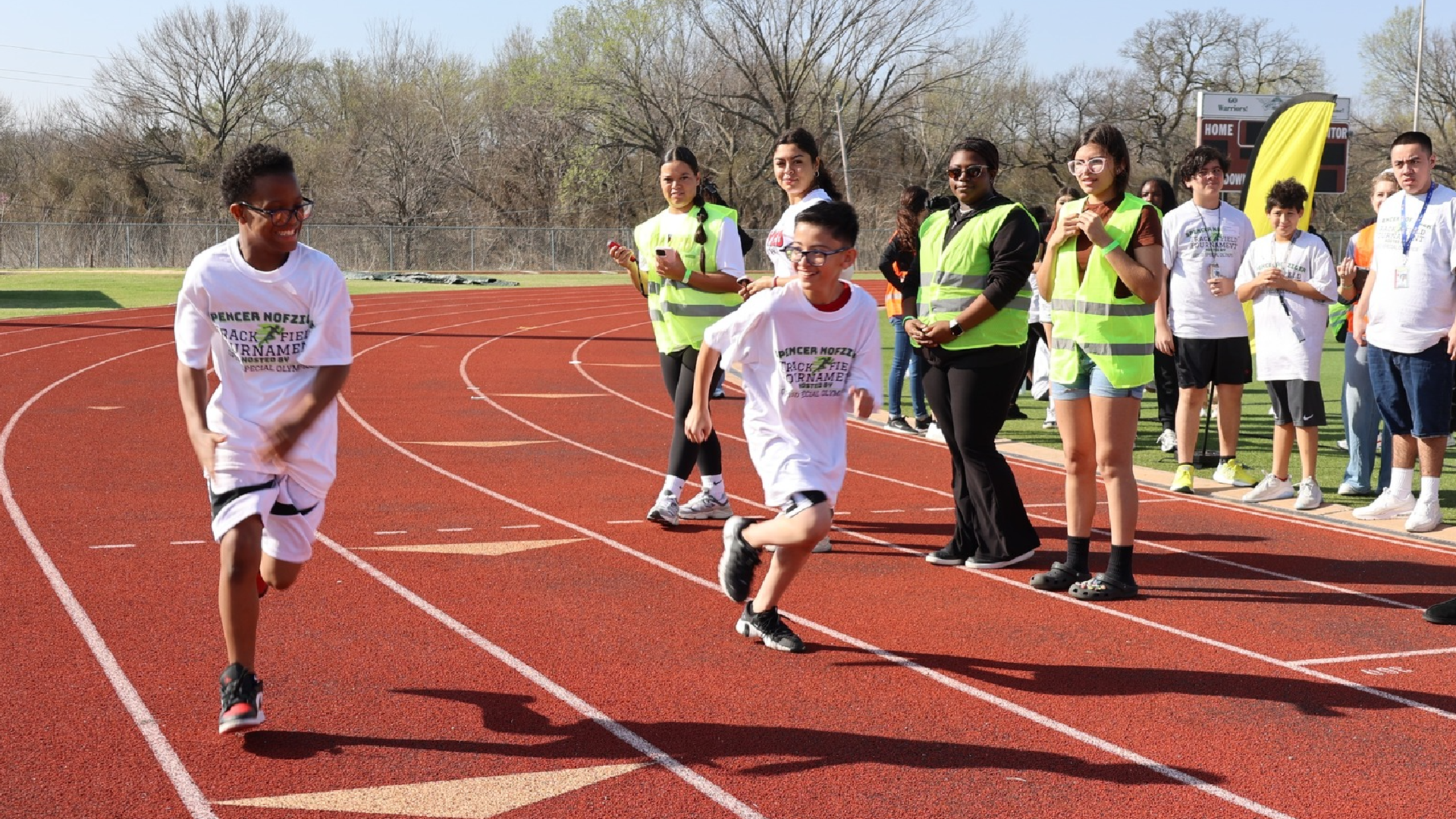 A group of people, some wearing bright yellow vests, are gathered on a running track, with several individuals running on the track in the foreground.