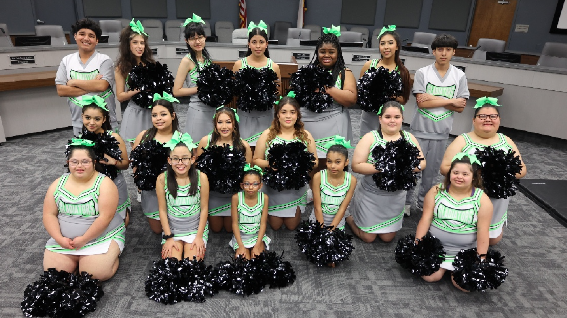 A group of young women in matching black and green cheerleader uniforms, with pom-poms, posing together in a room with a tiled floor and walls.