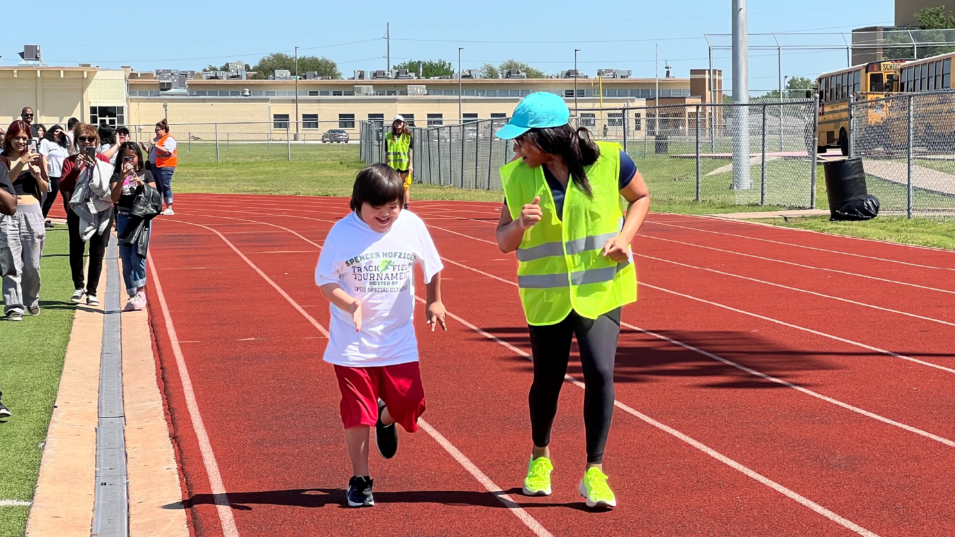 The image shows a group of people, including a person wearing a high-visibility vest, walking on a red running track surrounded by buildings and fencing.