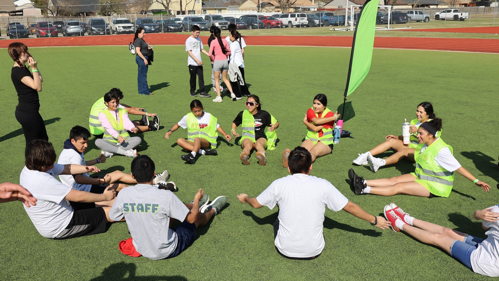A group of people, some wearing staff uniforms, are gathered on a green sports field, engaged in what appears to be a team-building or training activity.