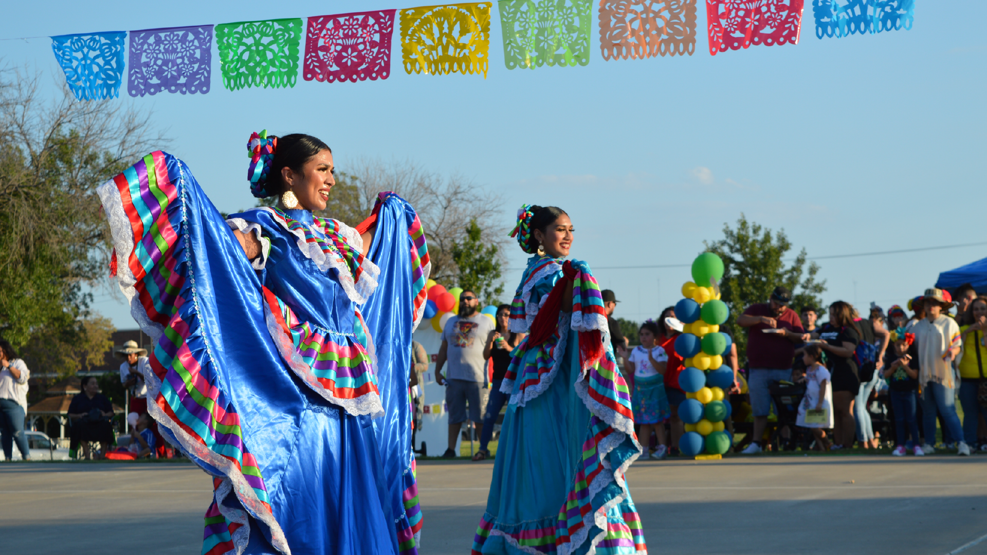 Colorful decorative flags hang overhead, while two individuals in vibrant, traditional-style clothing stand in the foreground against a crowd of people.