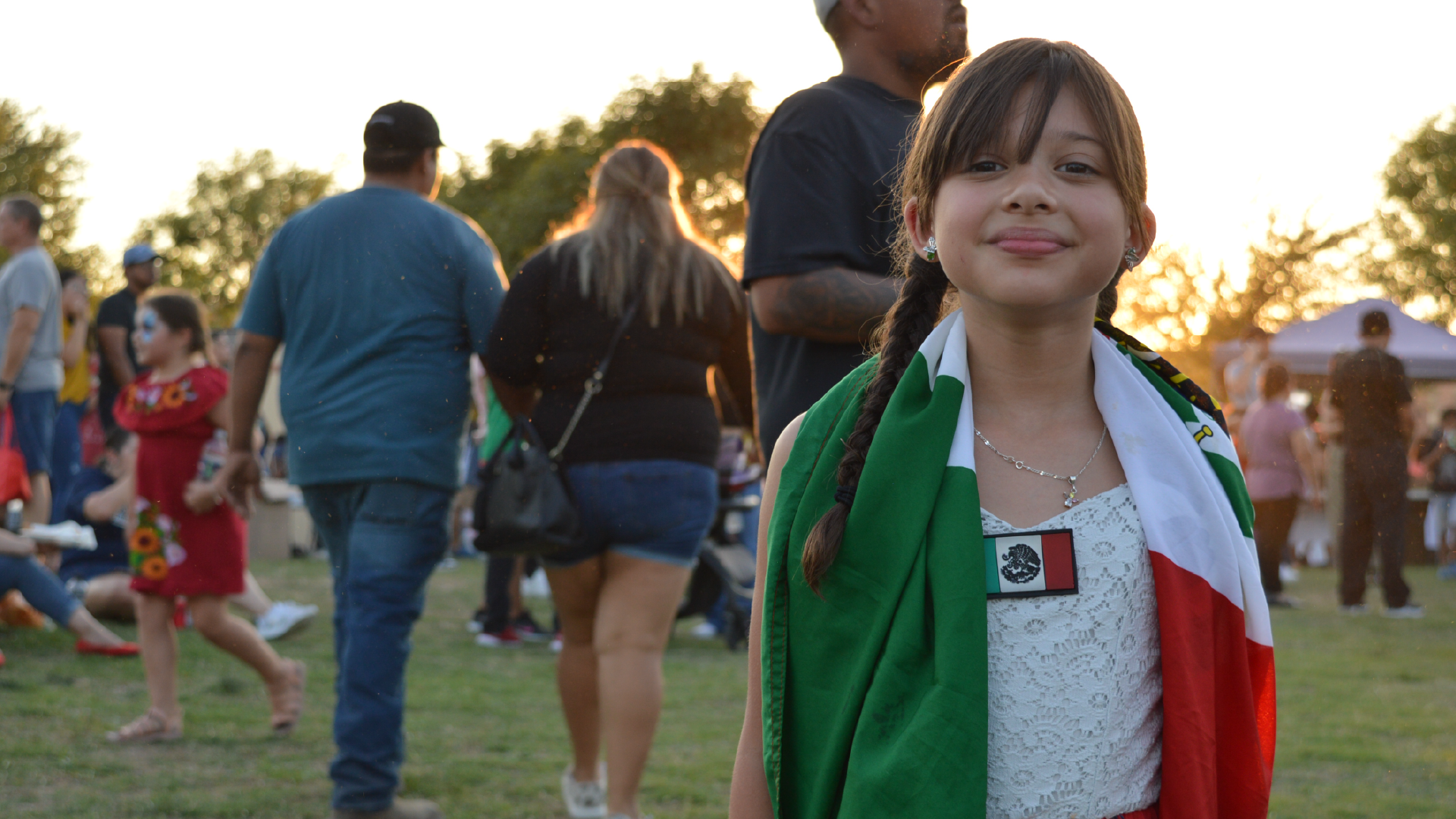 A young girl wearing a green, white, and red flag-like garment stands in the foreground, with a group of people visible in the background.