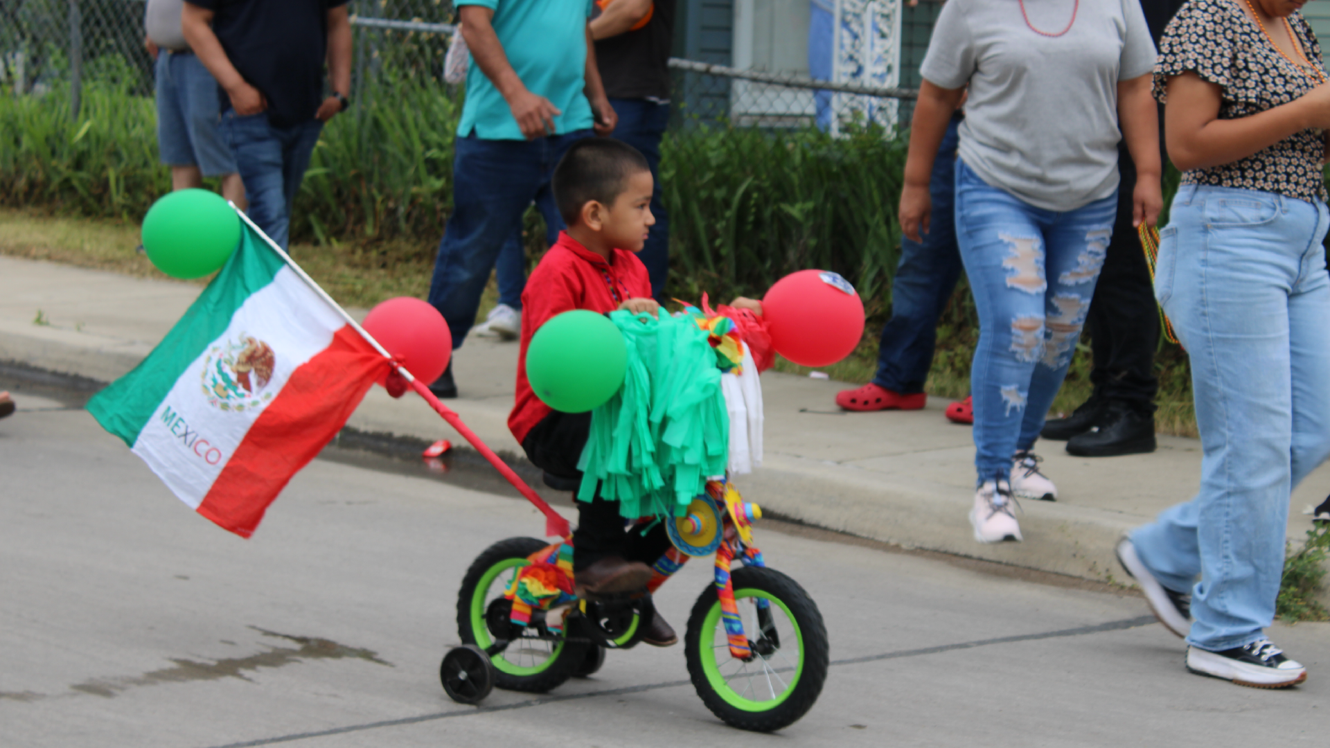 A young child riding a small bicycle decorated with Mexican flags and colorful balloons, surrounded by a group of adults standing in the background.