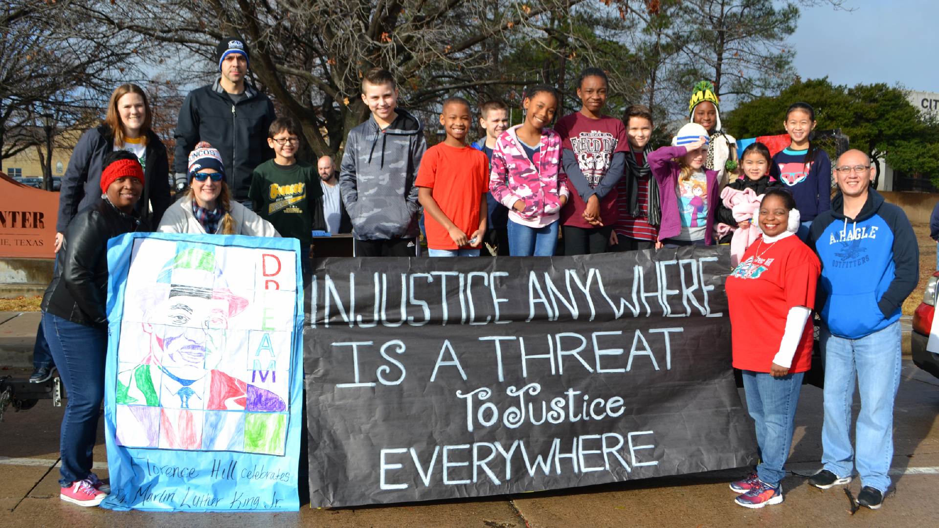 A group of people, including adults and children, are standing together holding a large sign that reads "Injustice anywhere is a threat to justice everywhere" in front of a building.