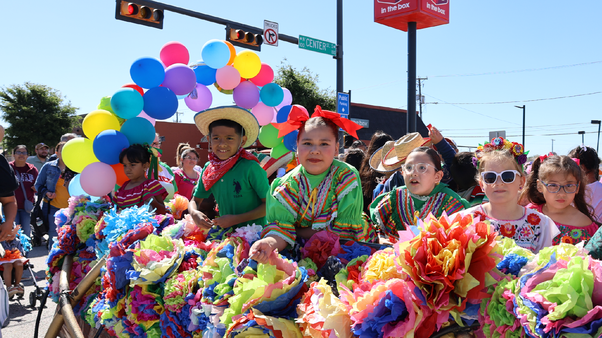 A colorful parade with people wearing vibrant costumes and carrying balloons of various shapes and sizes, set against a backdrop of buildings and street signs.