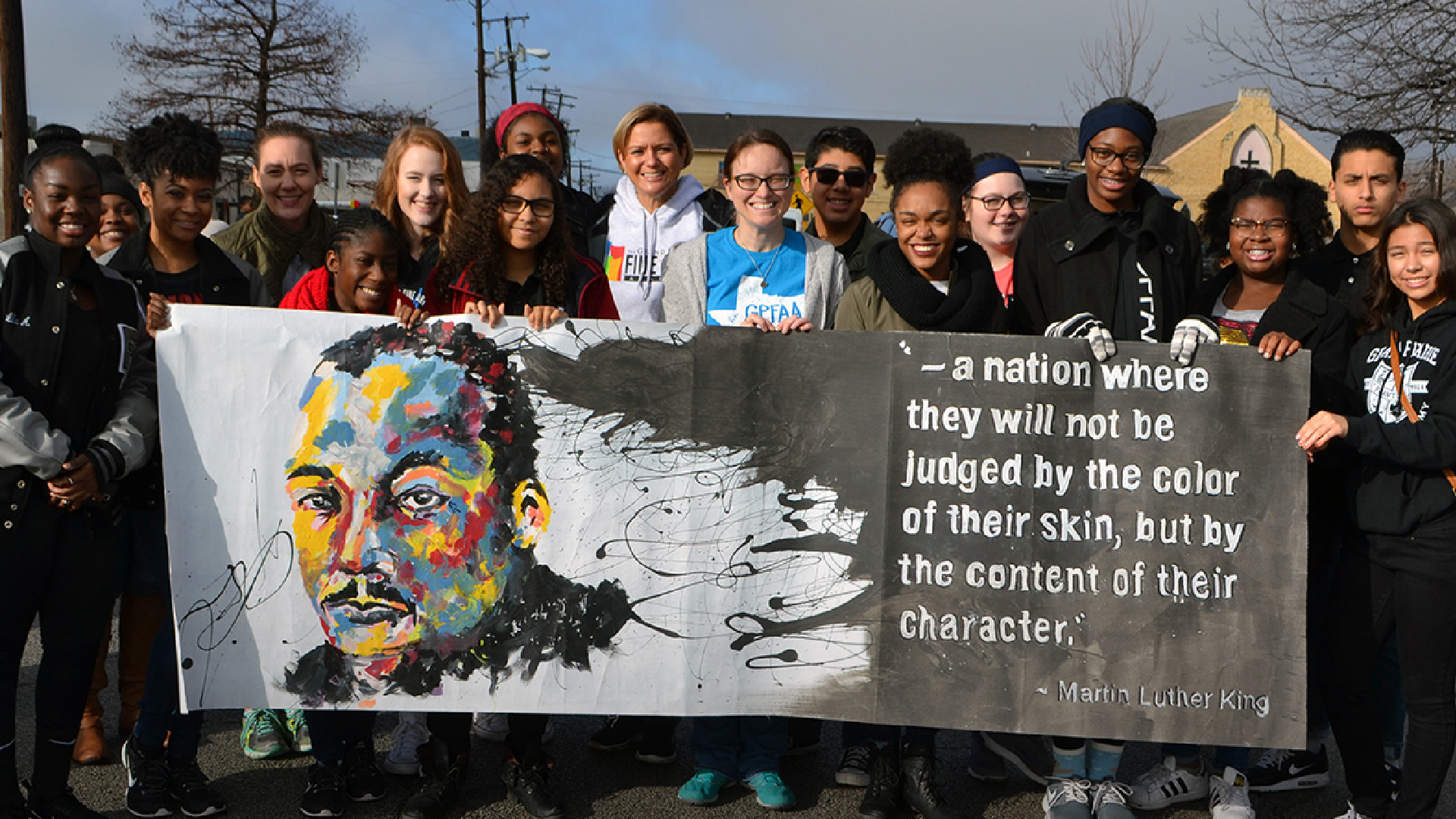 A group of people, including adults and children, are holding a large banner with a colorful portrait and a quote attributed to Martin Luther King Jr. about judging people by the content of their character rather than the color of their skin.