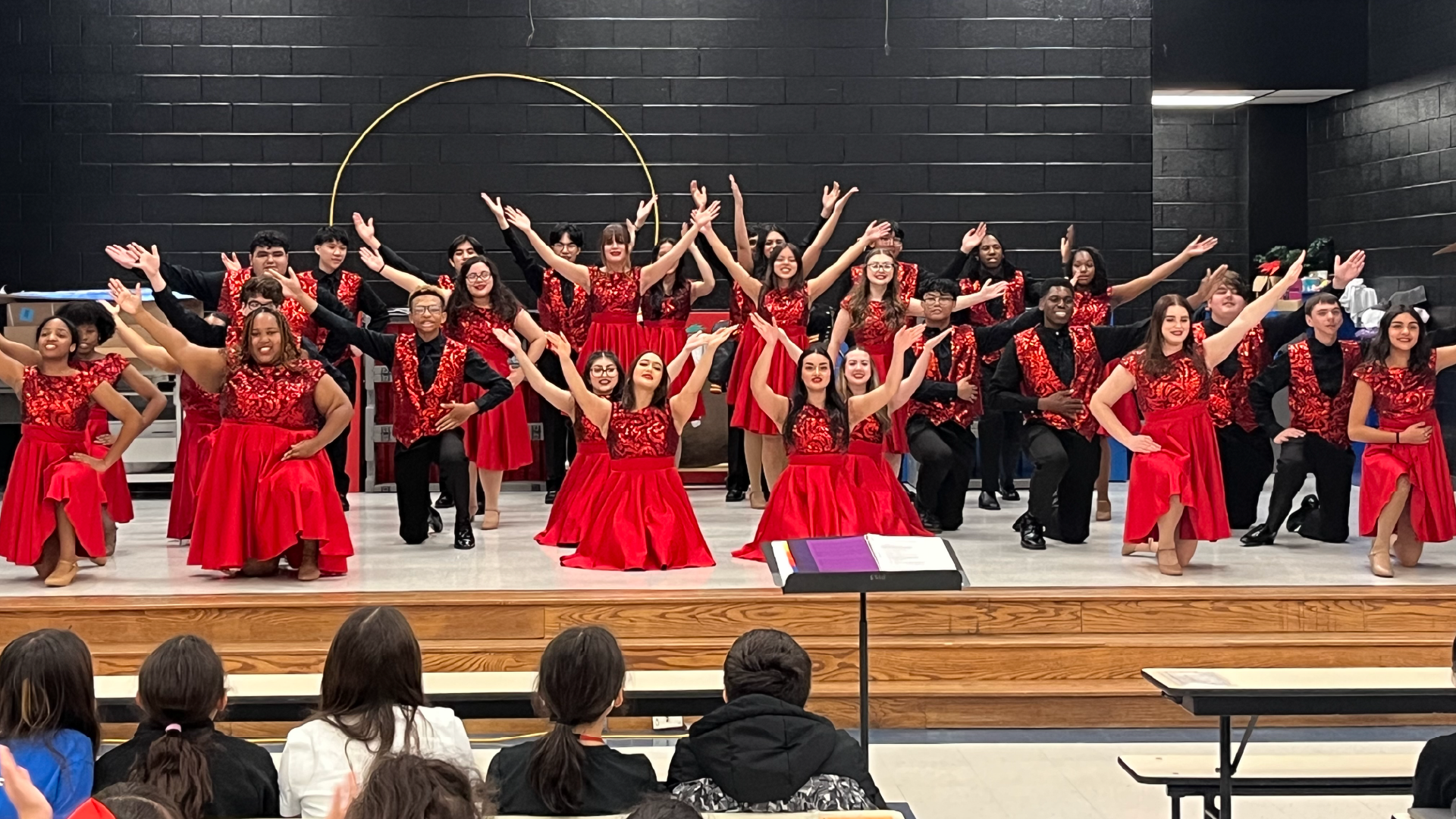 A large group of people in red dresses and costumes are performing on a stage, with a black brick wall in the background.