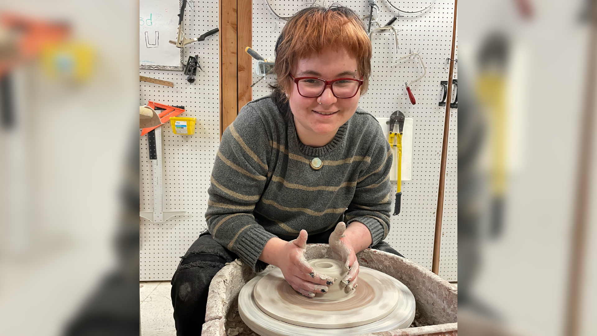 A person with glasses and reddish-brown hair is seated at a pottery wheel, intently focused on shaping a clay object in the foreground, with various tools and equipment visible in the background.