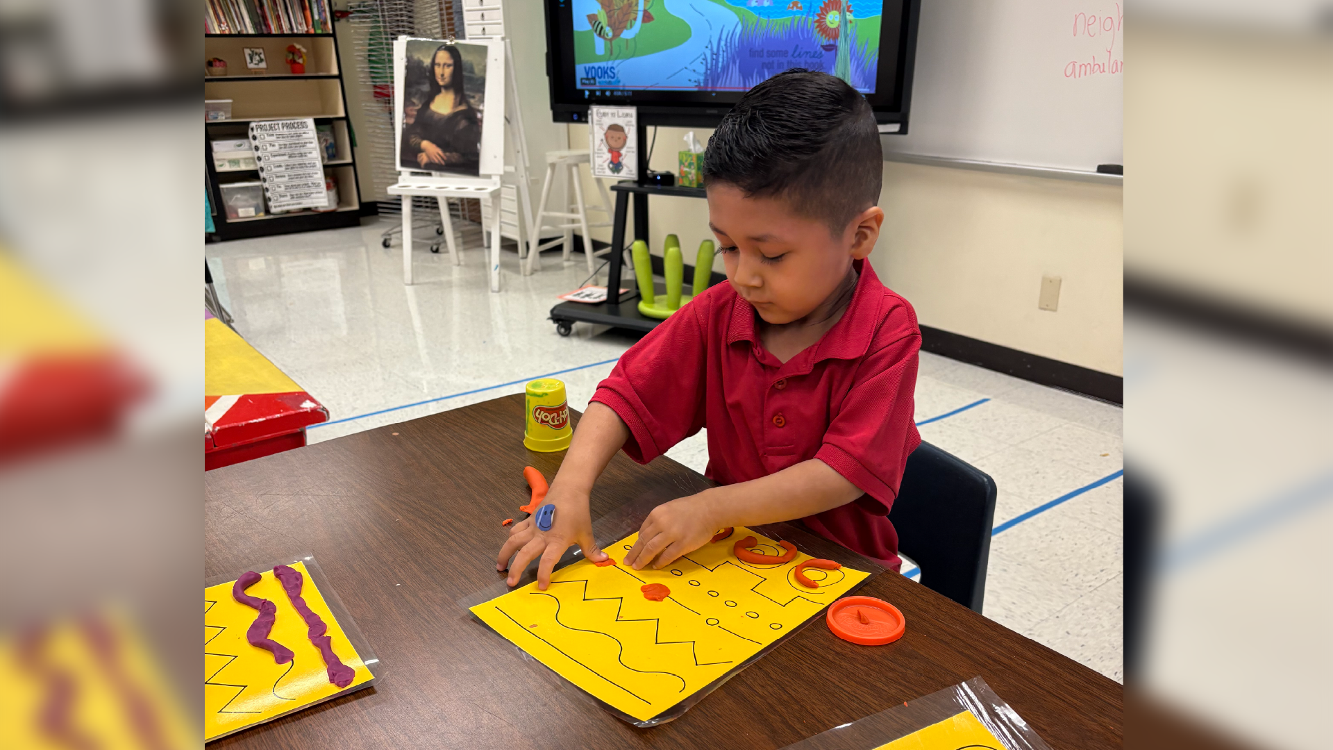A young boy in a red shirt is sitting at a table, intently working on a puzzle with colorful pieces in the foreground, while various framed artworks and a television screen can be seen in the background.