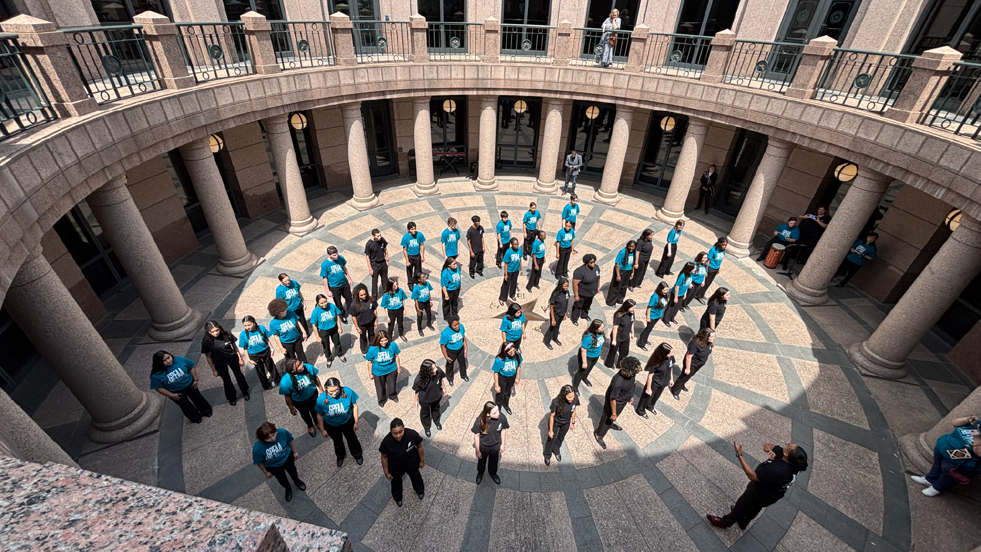 A large group of people in blue and black clothing standing in a circular formation within a grand, ornate architectural structure with arched entryways and columns.