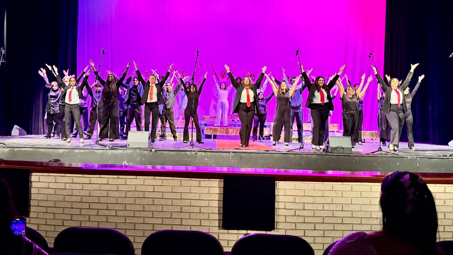 A group of people on a stage, dressed in formal attire, with their arms raised in celebration against a vibrant purple background.
