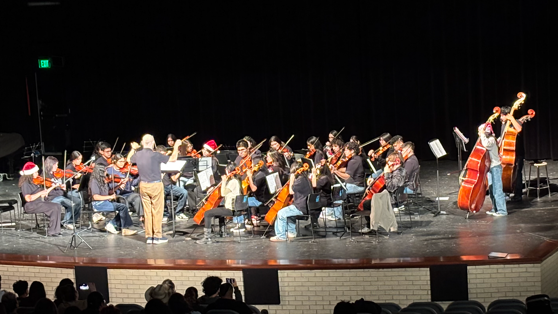 A large orchestra performing on a stage, with musicians playing various instruments and wearing colorful attire, while an audience is visible in the background.