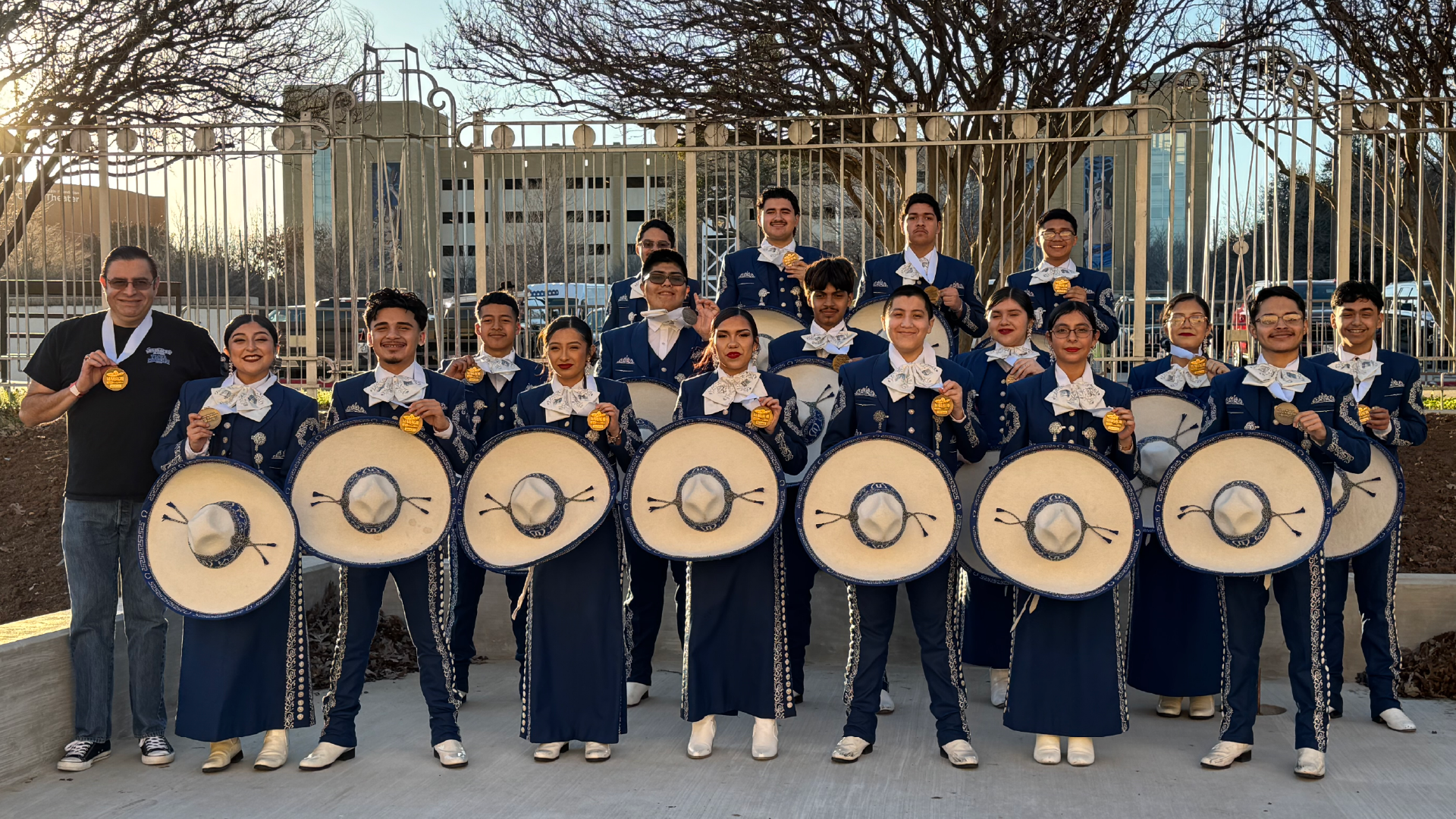 A group of young people in matching uniforms, holding large round instruments, stand in front of a metal gate structure in an outdoor setting with trees in the background.