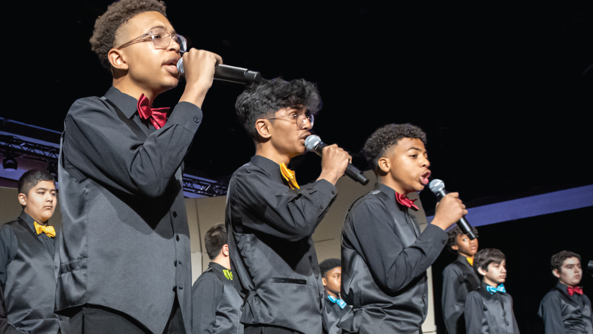 A group of young performers in formal attire singing into microphones on a stage with colorful lighting in the background.