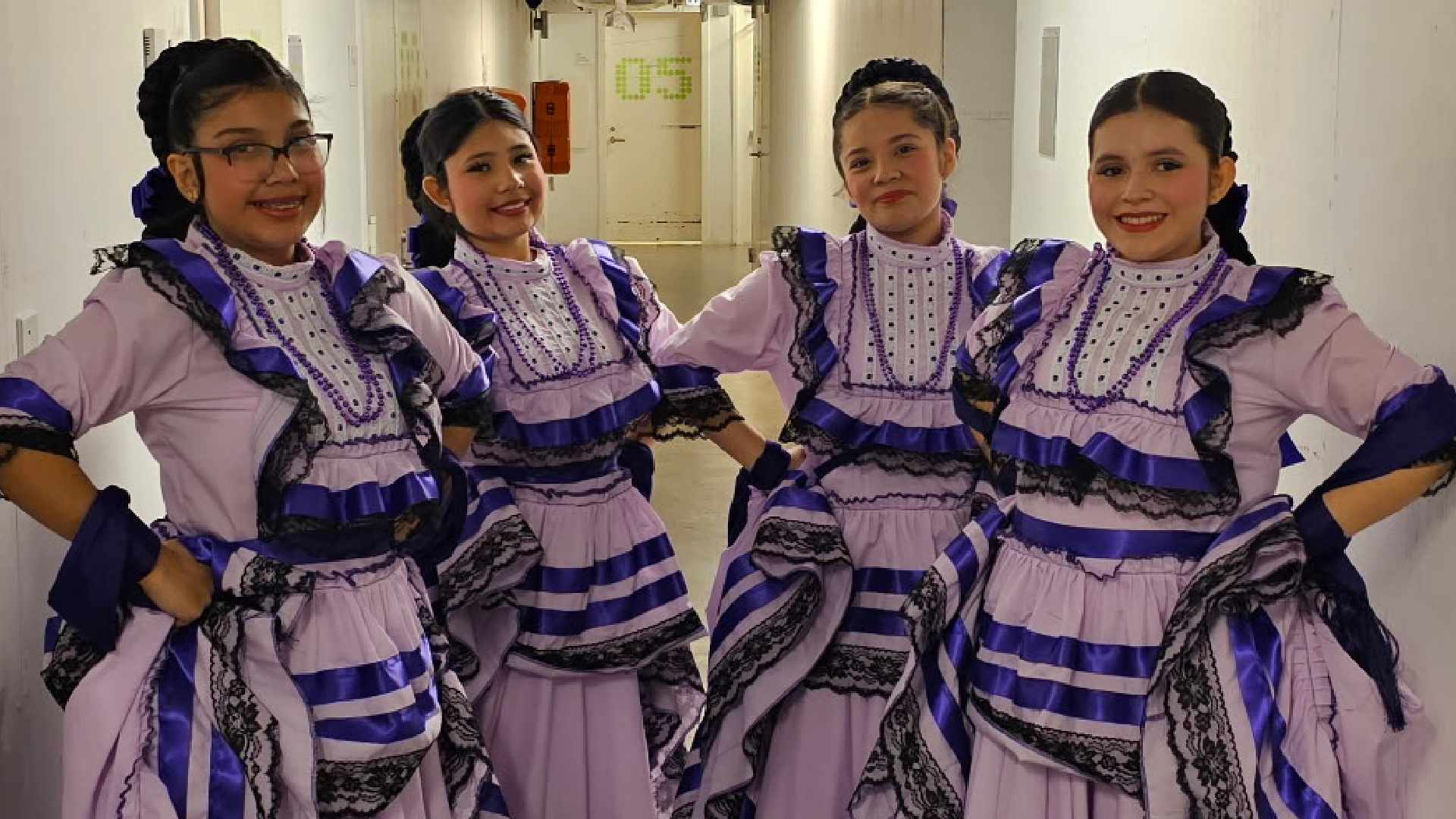 Four young women wearing traditional purple and white dresses with intricate patterns and ruffles, posing together in what appears to be a hallway or indoor setting.
