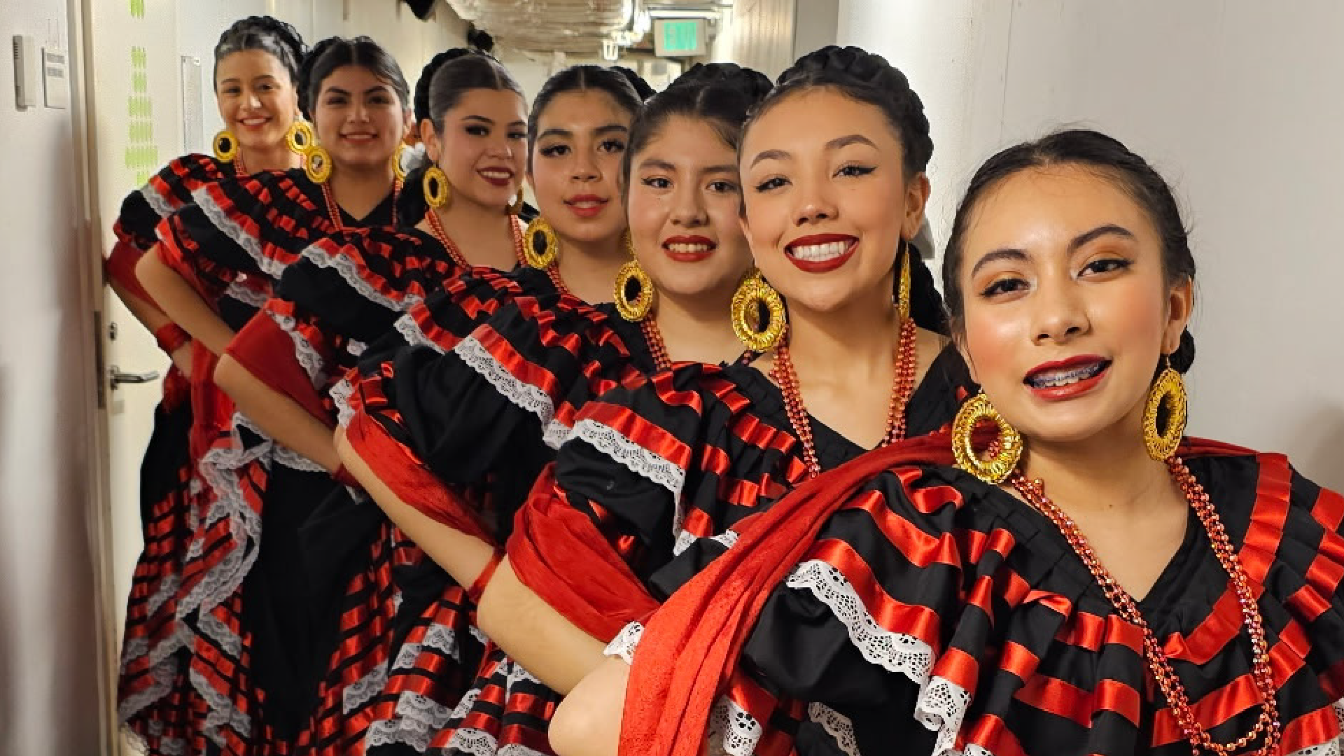 A group of women wearing vibrant, patterned dresses and large, dangling earrings stand together in what appears to be a hallway or indoor setting.