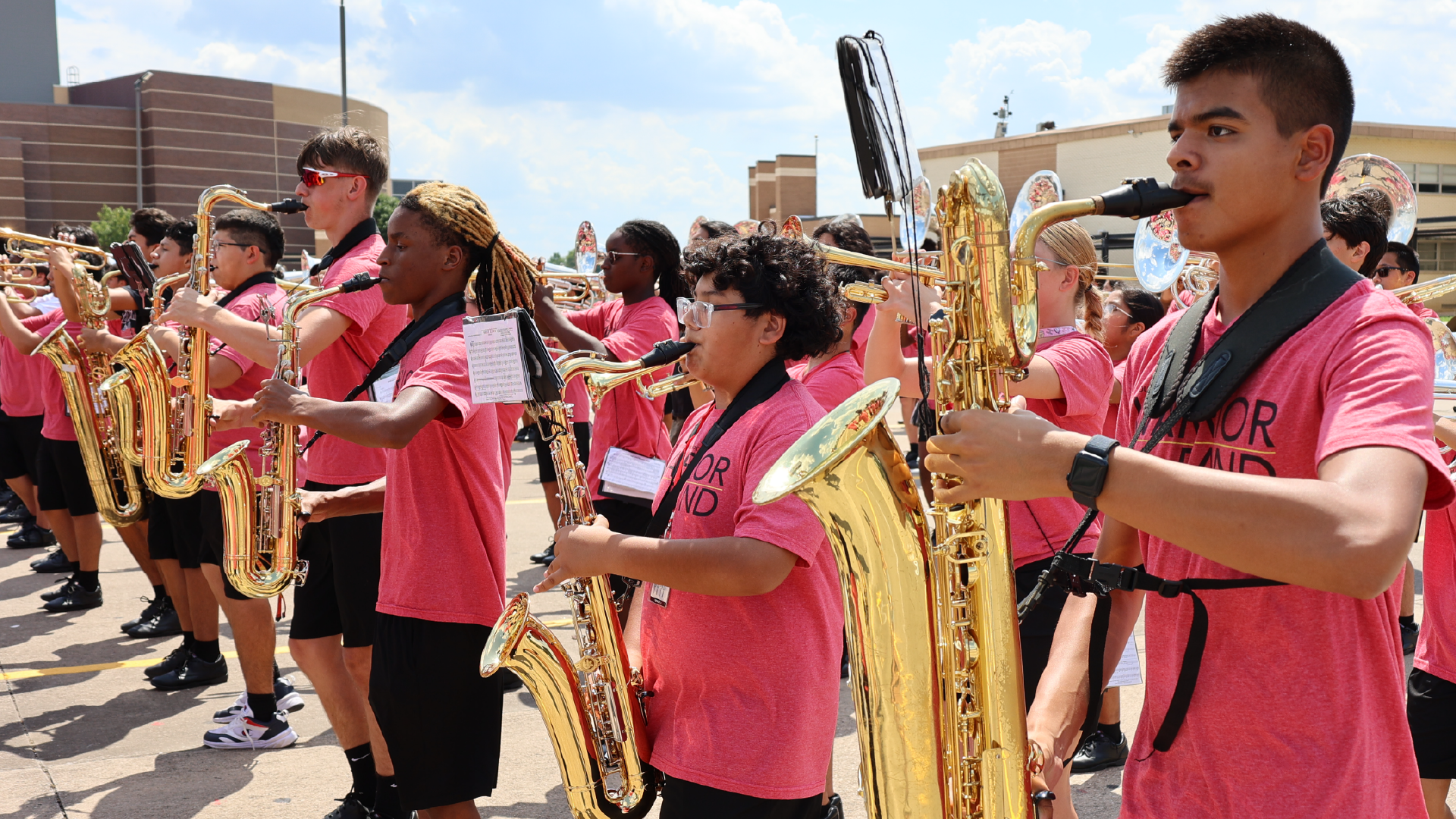 A group of people wearing matching pink shirts and holding various brass instruments, standing in front of a building with a cloudy sky in the background.