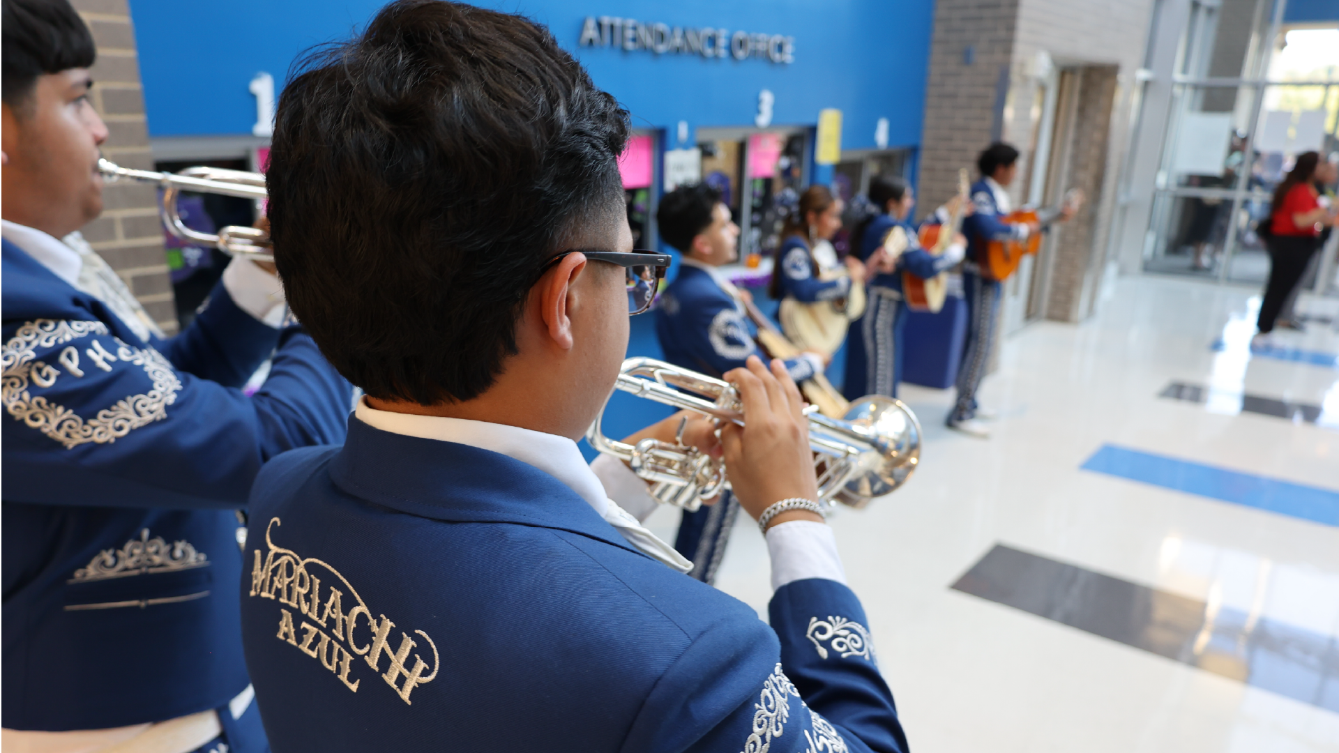 The image shows a person in a blue uniform with gold embroidery, standing in a crowded hallway with a blue wall and various signs and displays in the background.